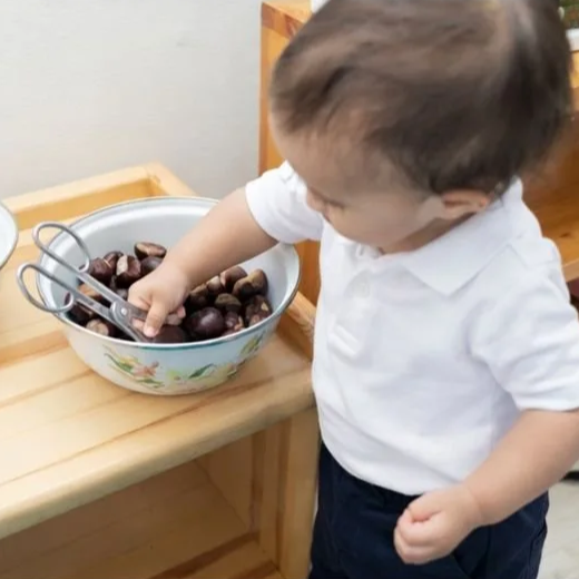 A young child wearing a white shirt and dark pants reaching into a bowl of chestnuts with tongs.