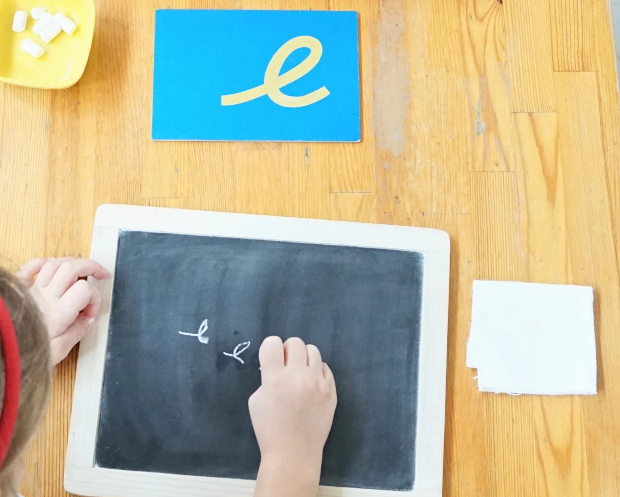 Child writing the letter 'e' on a small black chalkboard with a hand, with letter 'e' and a learning card showing the cursive 'e' on a blue background on a wooden table.