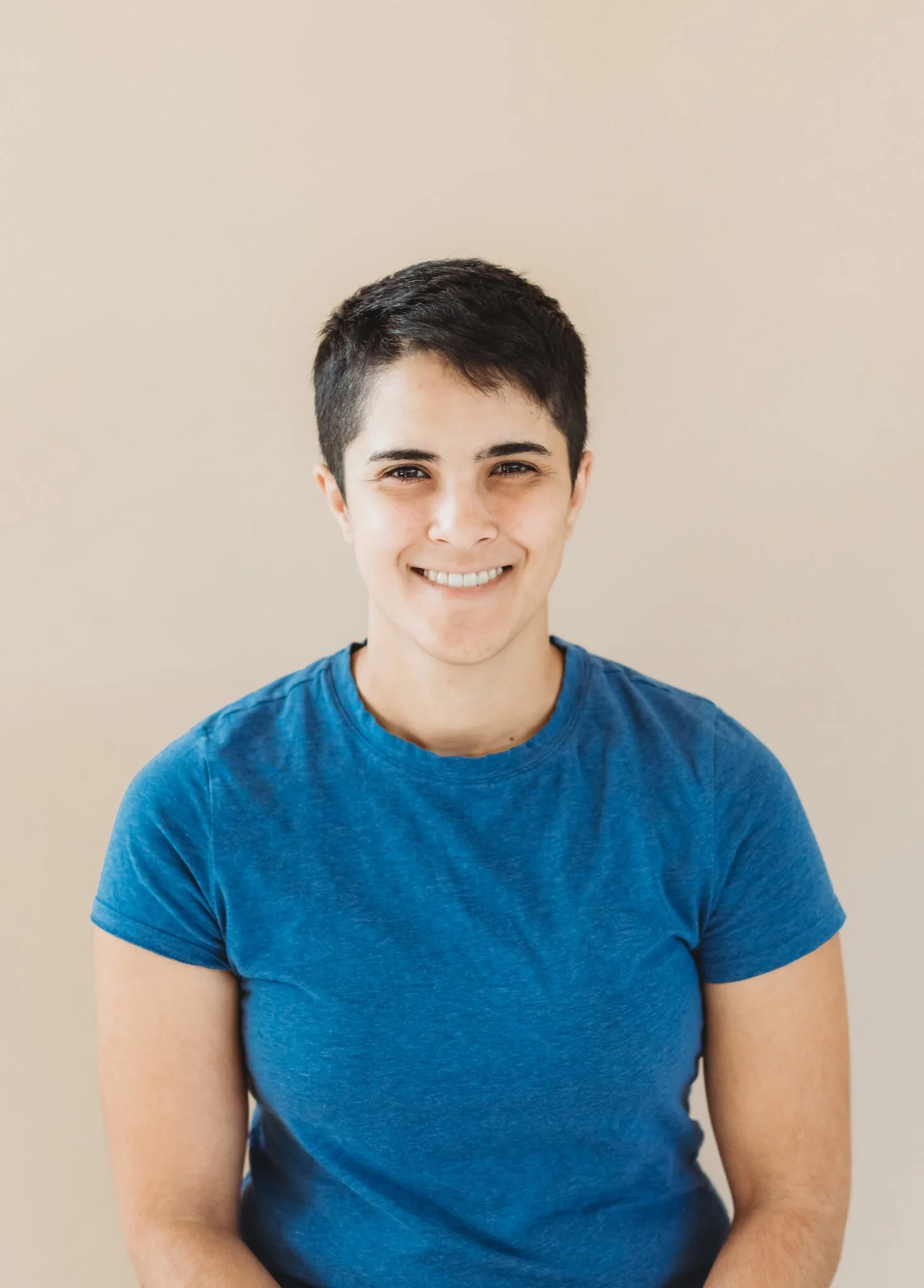 A person with short dark hair, wearing a blue t-shirt, smiling and looking at the camera, against a plain beige background.