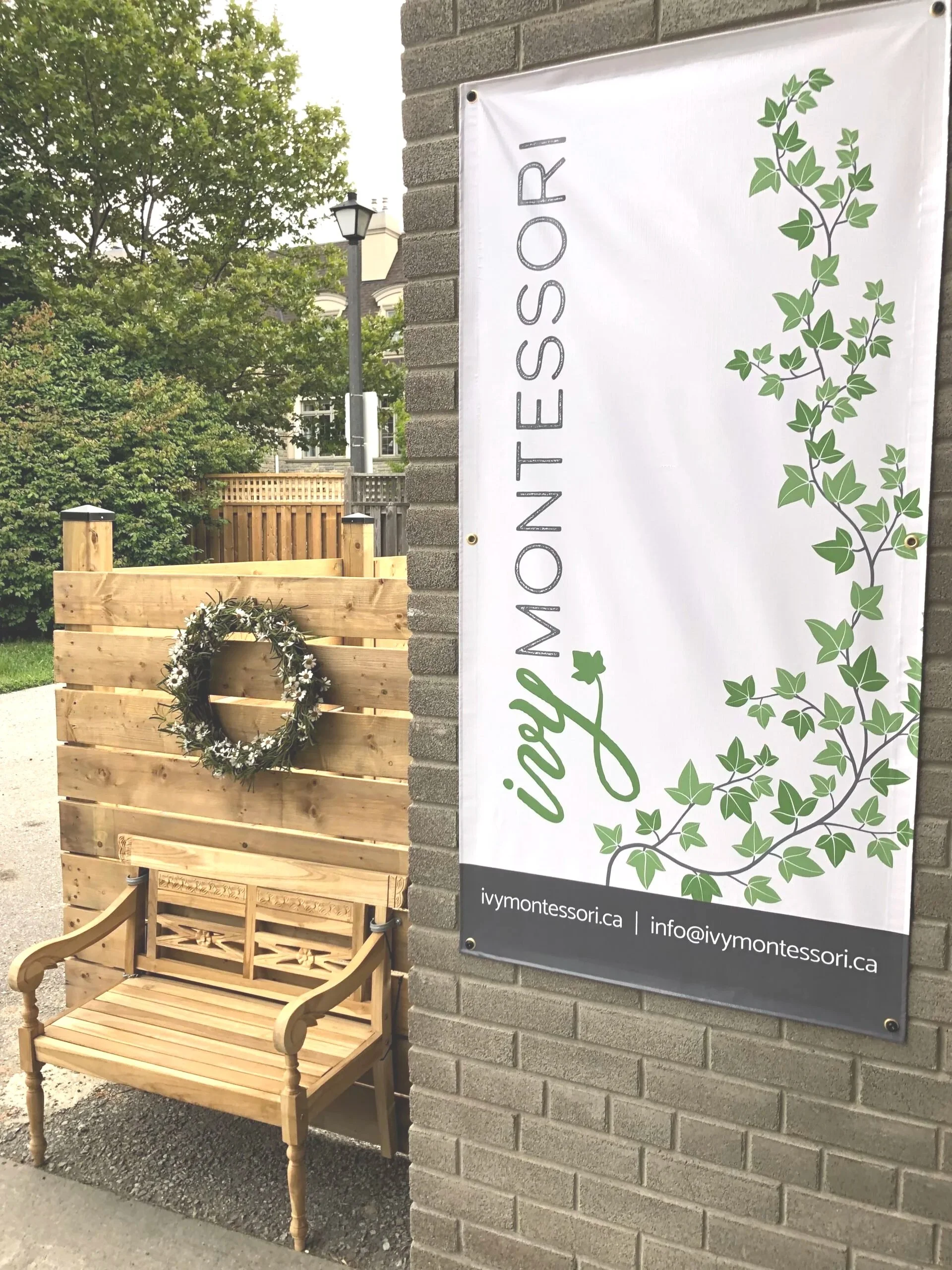 Sign with text 'iYMONTESSORI' and plant vine design, attached to a brick wall, with a wooden bench with armrests and a floral wreath in front of the wall.