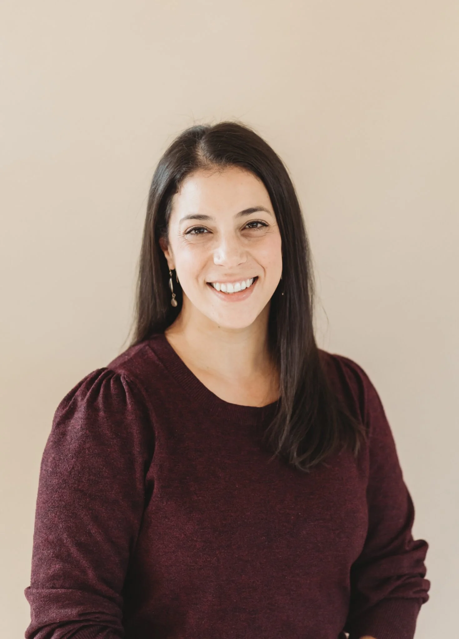 A woman with long dark hair, smiling, wearing a burgundy sweater and earrings, standing against a plain beige background.