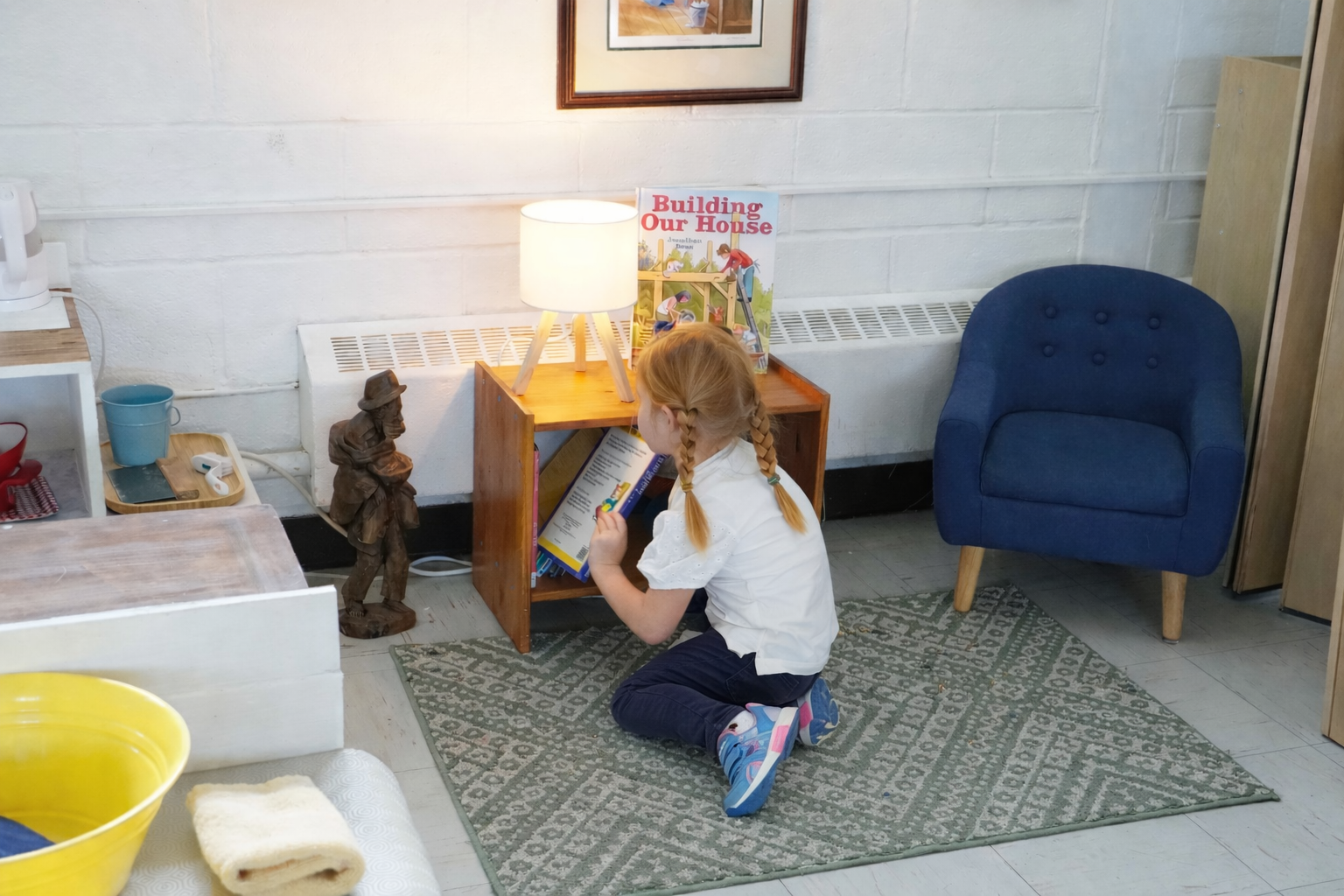 A young girl with blonde braided hair, wearing a white shirt and dark pants, kneeling on a patterned rug, looking at a book under a small wooden side table with a lamp on top. There is a blue armchair nearby and a framed picture on the wall behind.