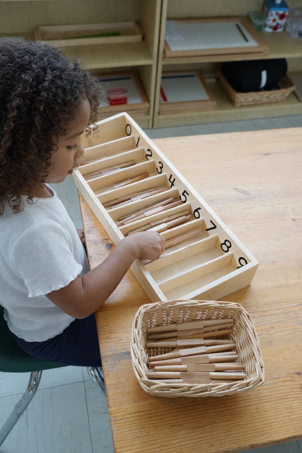A young girl with curly hair arranging wooden sticks in a numbered Montessori desk with compartments, with a basket of more sticks nearby, in a classroom setting.