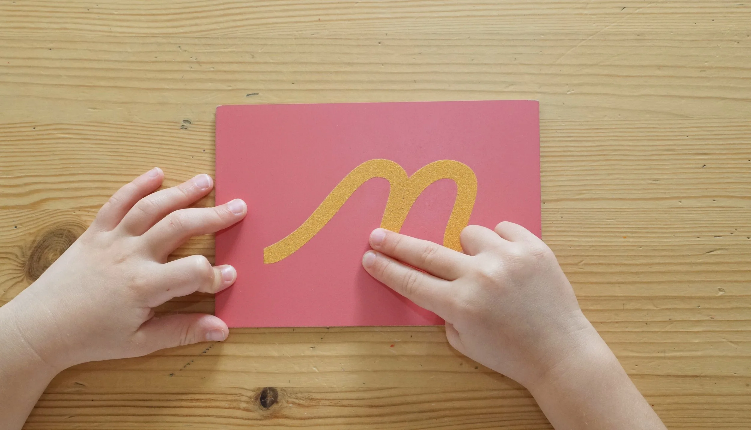 A child's hands placing a gold glittery smiley face sticker on a pink card on a wooden table.
