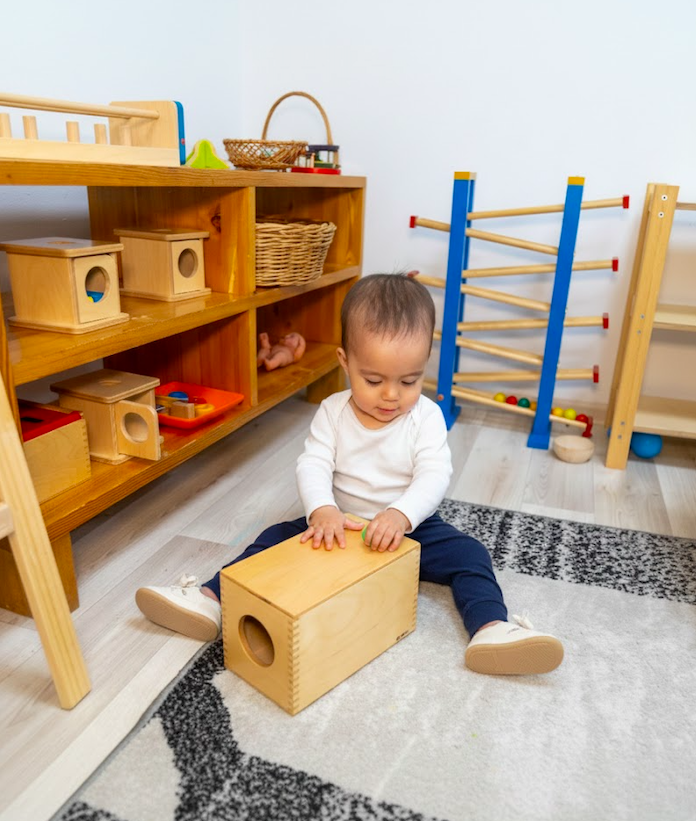 A toddler sitting on a rug with a wooden toy box on their lap in a playroom with shelves and climbing equipment