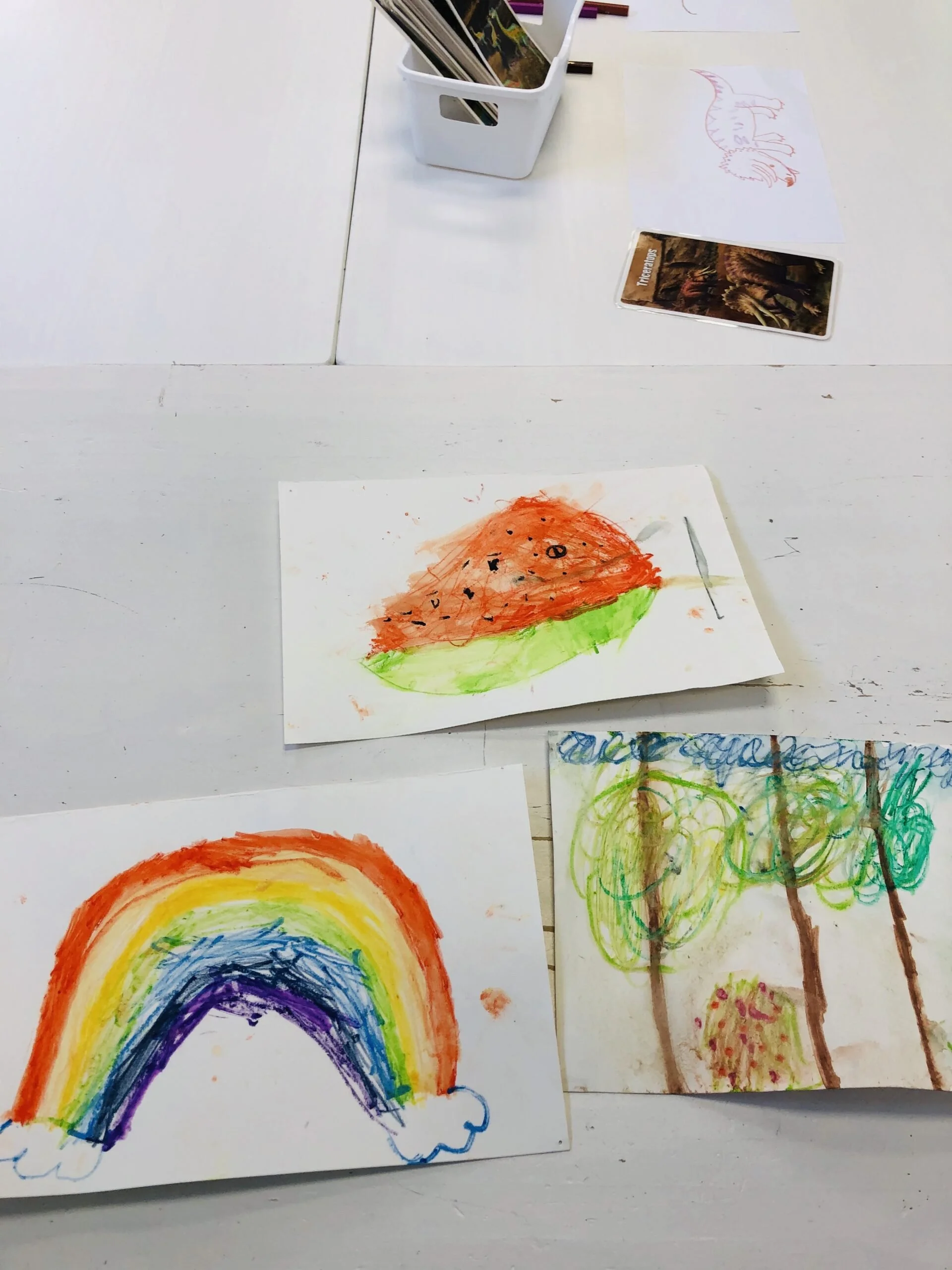 Child's colorful drawings of a rainbow, a watermelon, and trees on white paper, placed on a white table.