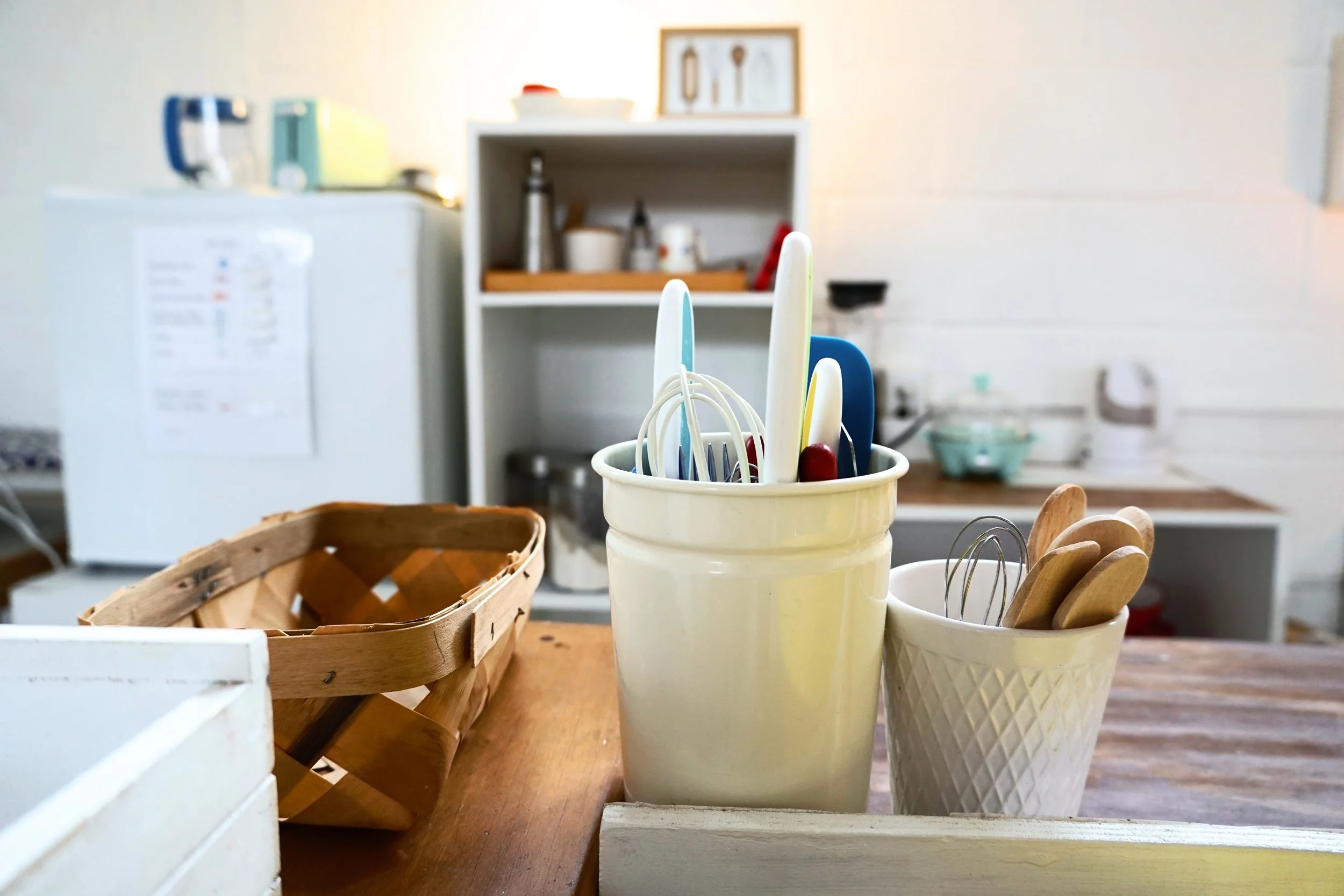 Kitchen with utensils in baskets on a wooden table, fridge in the background.