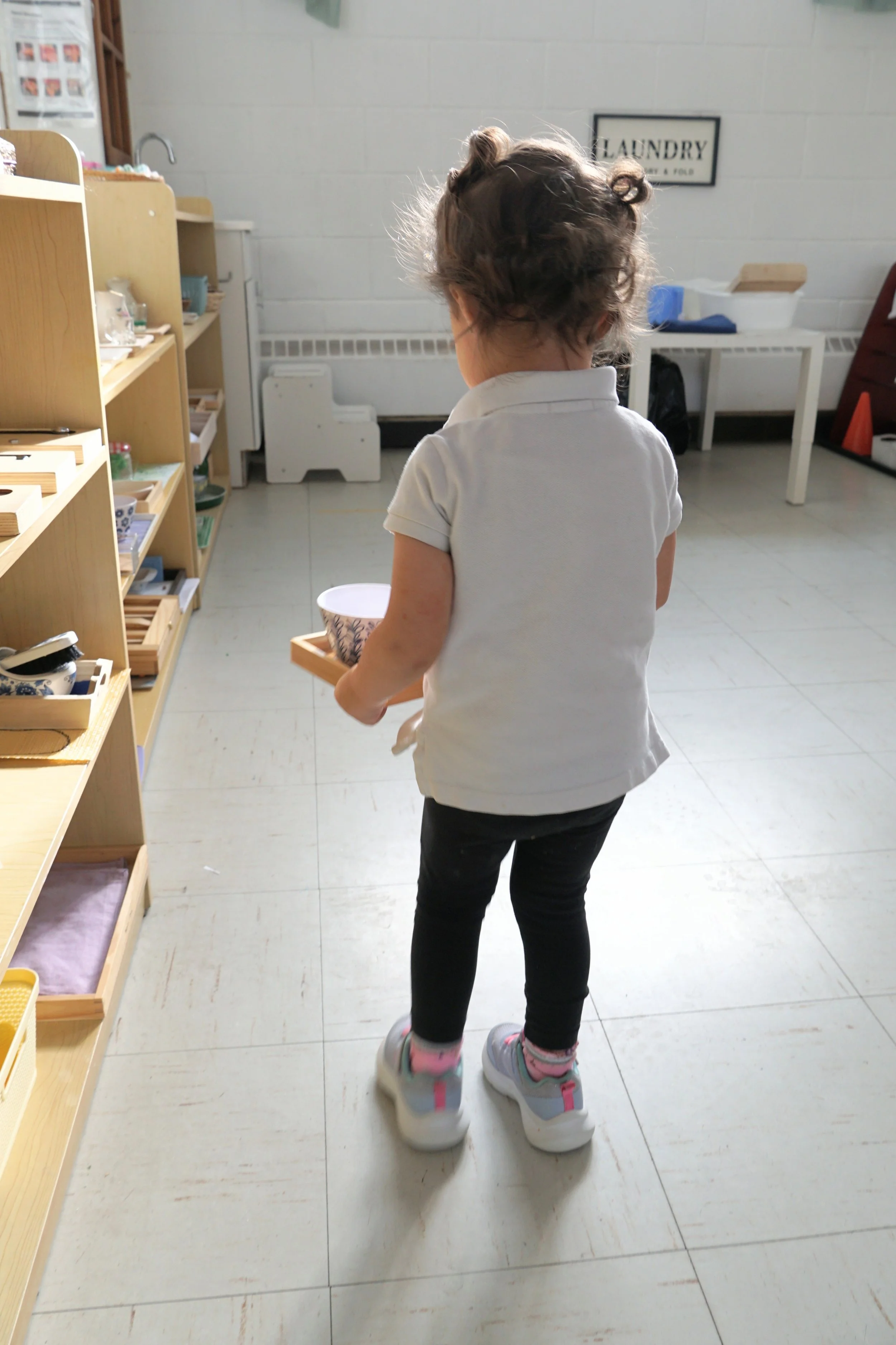 A young girl with curly hair wearing a white polo shirt, black pants, and sneakers, standing in a room with shelves of small items, holding a bowl in her right hand, near a sign that says 'Laundry' on the wall.