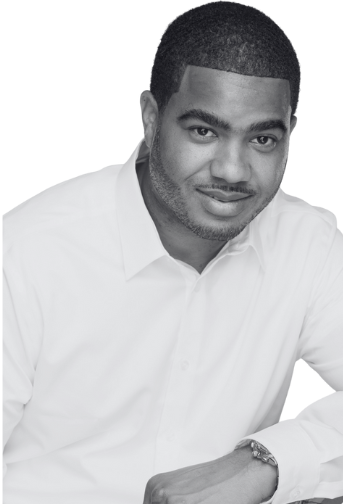 Black and white portrait of a man with short dark hair, a beard, and wearing a T-shirt and necklace, looking directly at the camera.