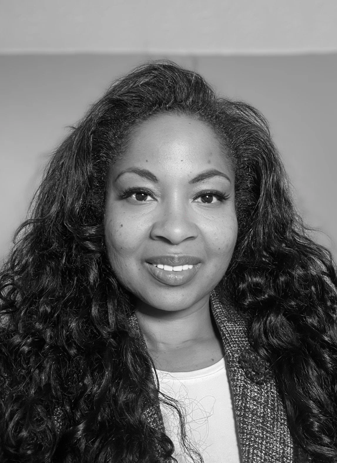 A woman with braided hair smiling, wearing a black turtleneck and pearl-embellished shoulder straps in a black and white photo.