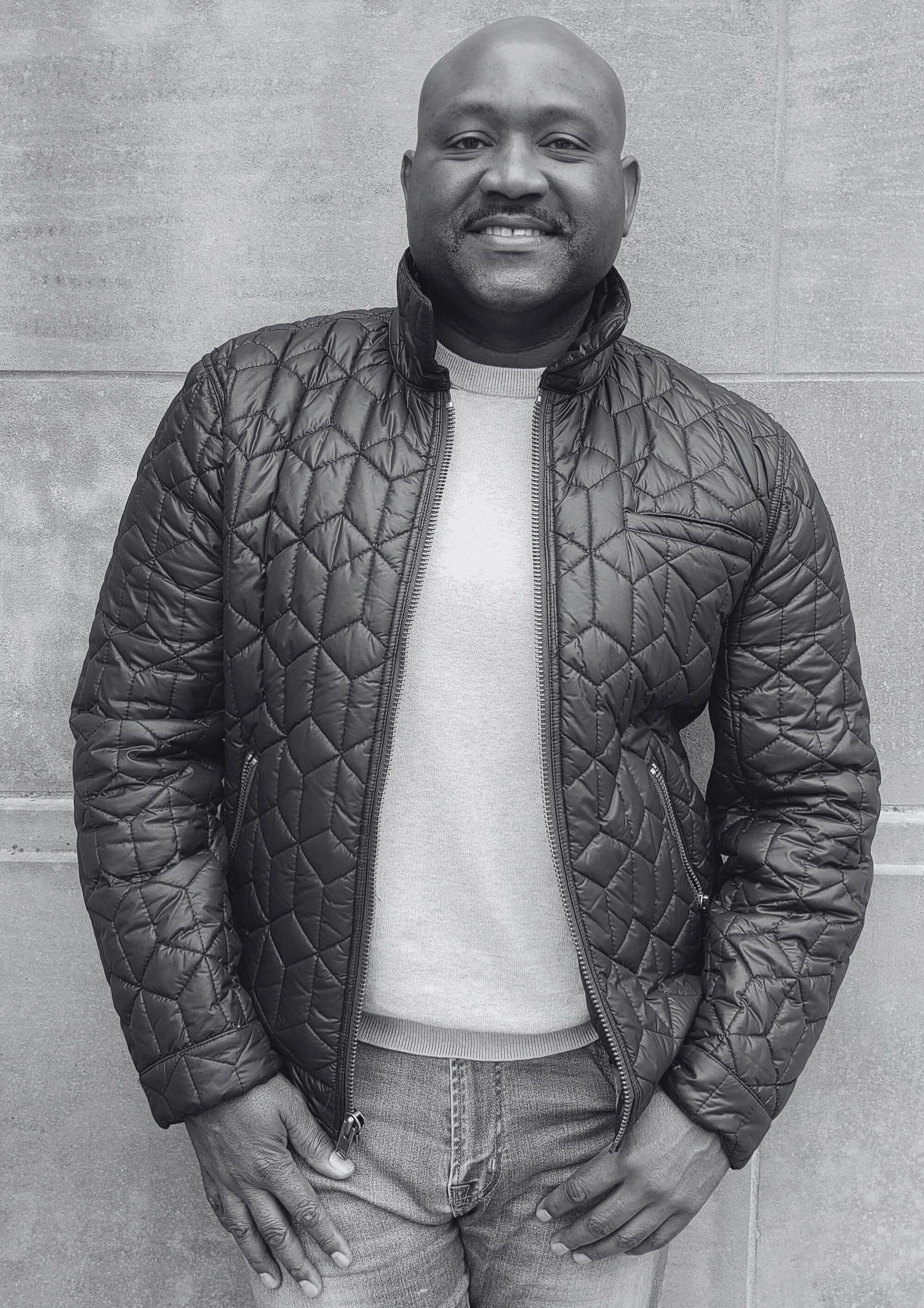 A smiling man in a quilted black jacket and jeans standing against a concrete wall.
