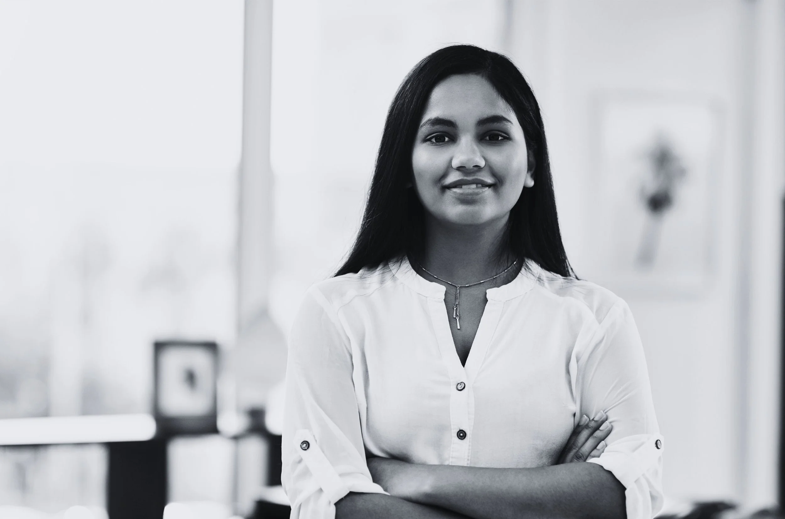 A smiling woman with long dark hair, wearing a white blouse and a necklace, standing indoors with crossed arms.