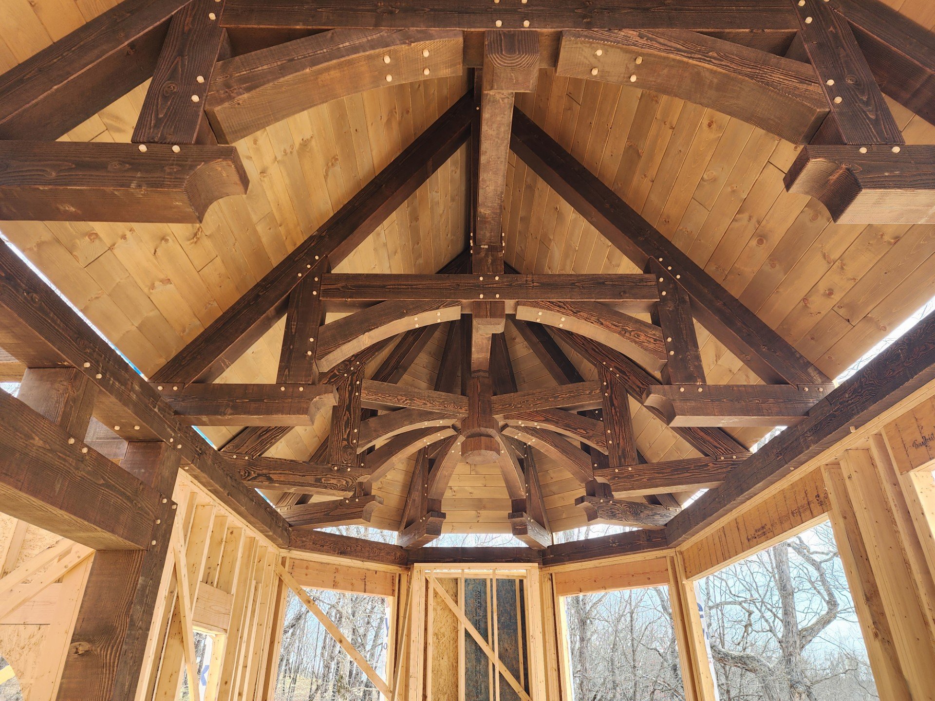 Looking up at the wooden framework of a building under construction, showing the ceiling with angled beams and exposed rafters, with partly finished walls and a view of trees outside through large window openings.