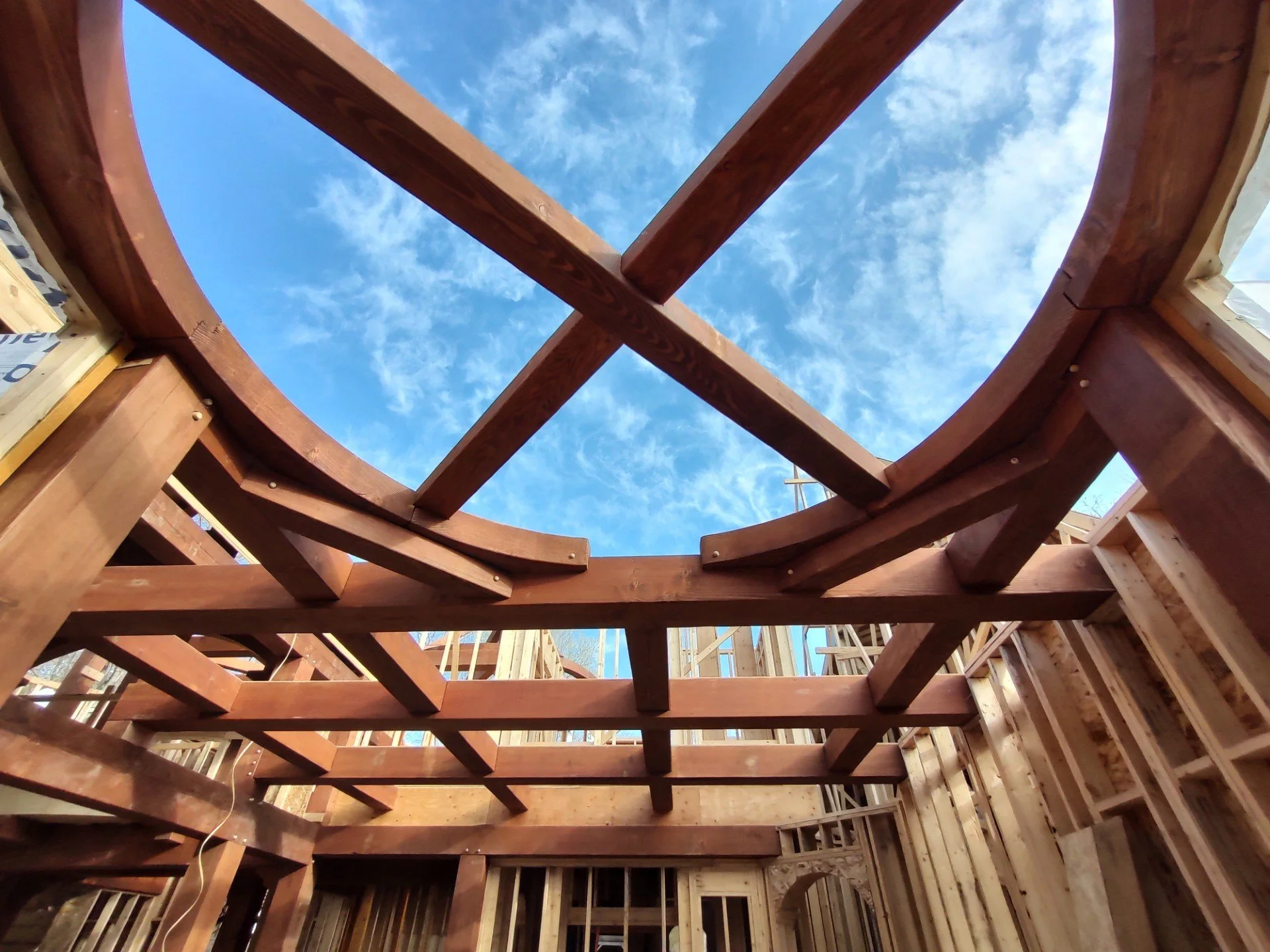 Looking up at the wooden framework of a building under construction, with blue sky and clouds visible through the open roof.