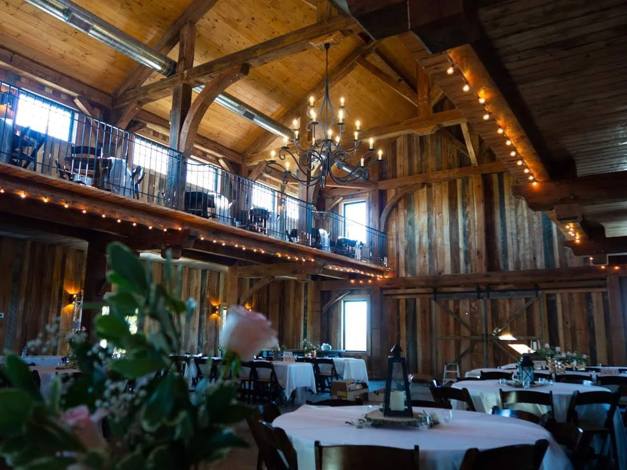 Interior of a rustic wooden event hall with high ceilings, a chandelier, string lights, and decorated tables for a celebration.