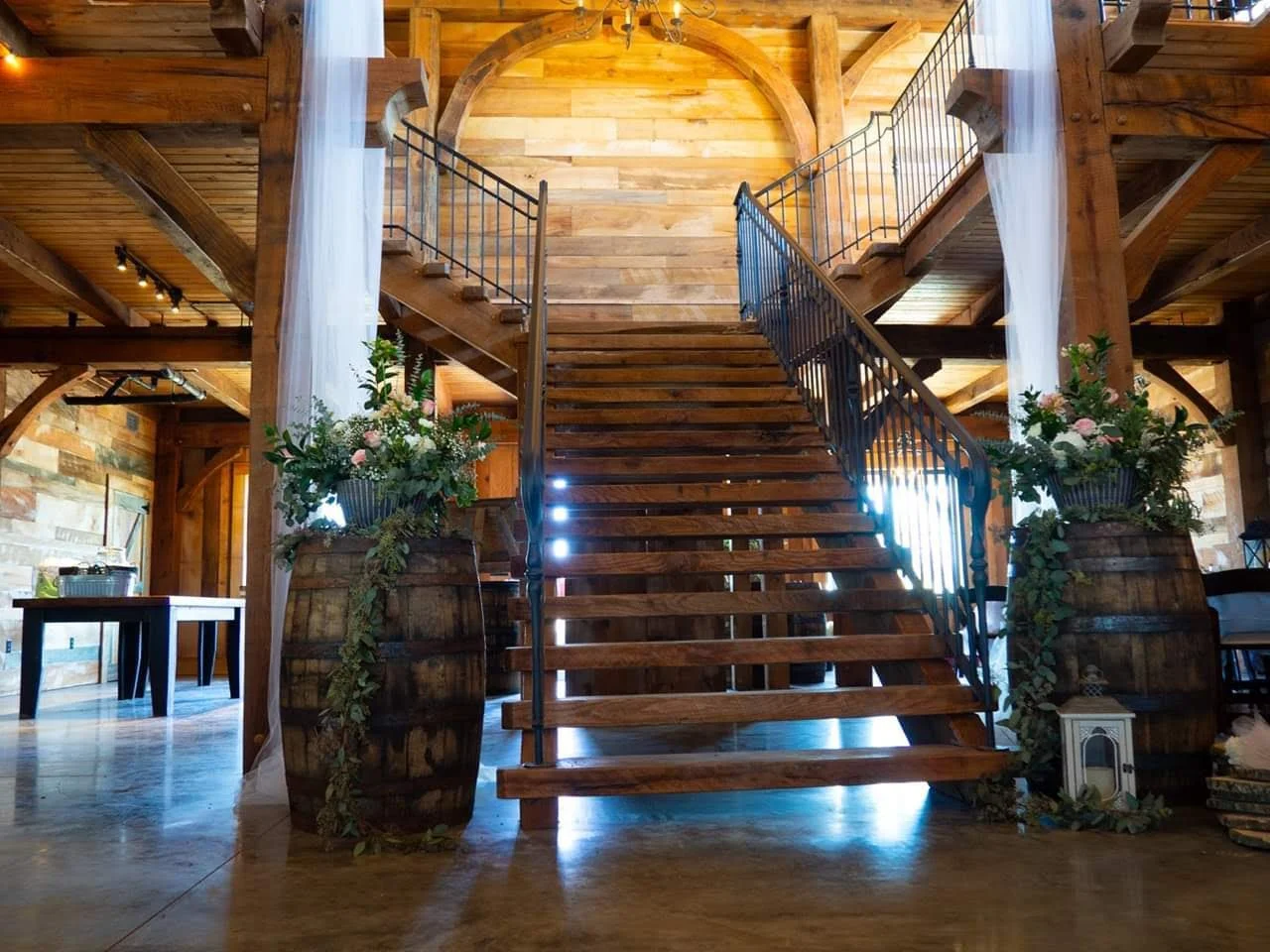 Wooden staircase leading to upper level in rustic interior with large flower arrangements in barrels on either side