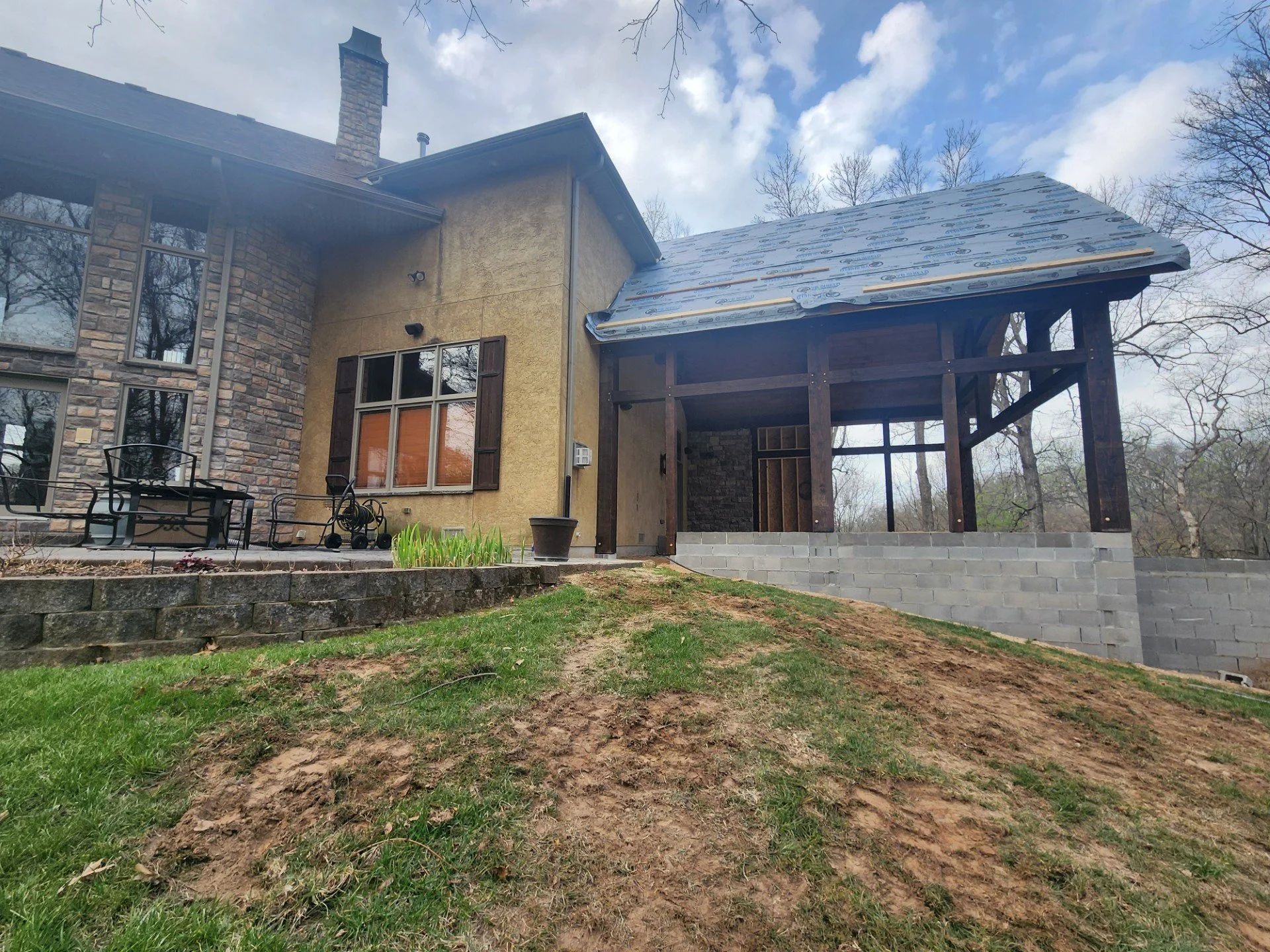 A house under construction with an addition being built. The house has stone and stucco exterior walls, multiple windows with shutters, and a chimney. The addition has a roof with roofing underlayment and wooden framing, with gray cinder block founda