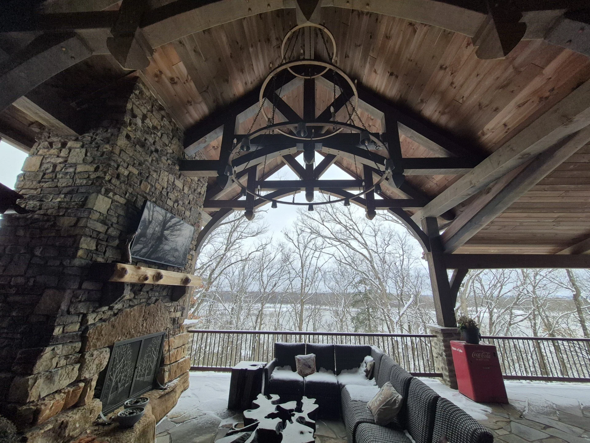 A covered outdoor patio with a stone fireplace, wooden ceiling, and large black chandelier. There are outdoor couches with pillows, a snow-covered table, and a view of snow-covered trees outside.