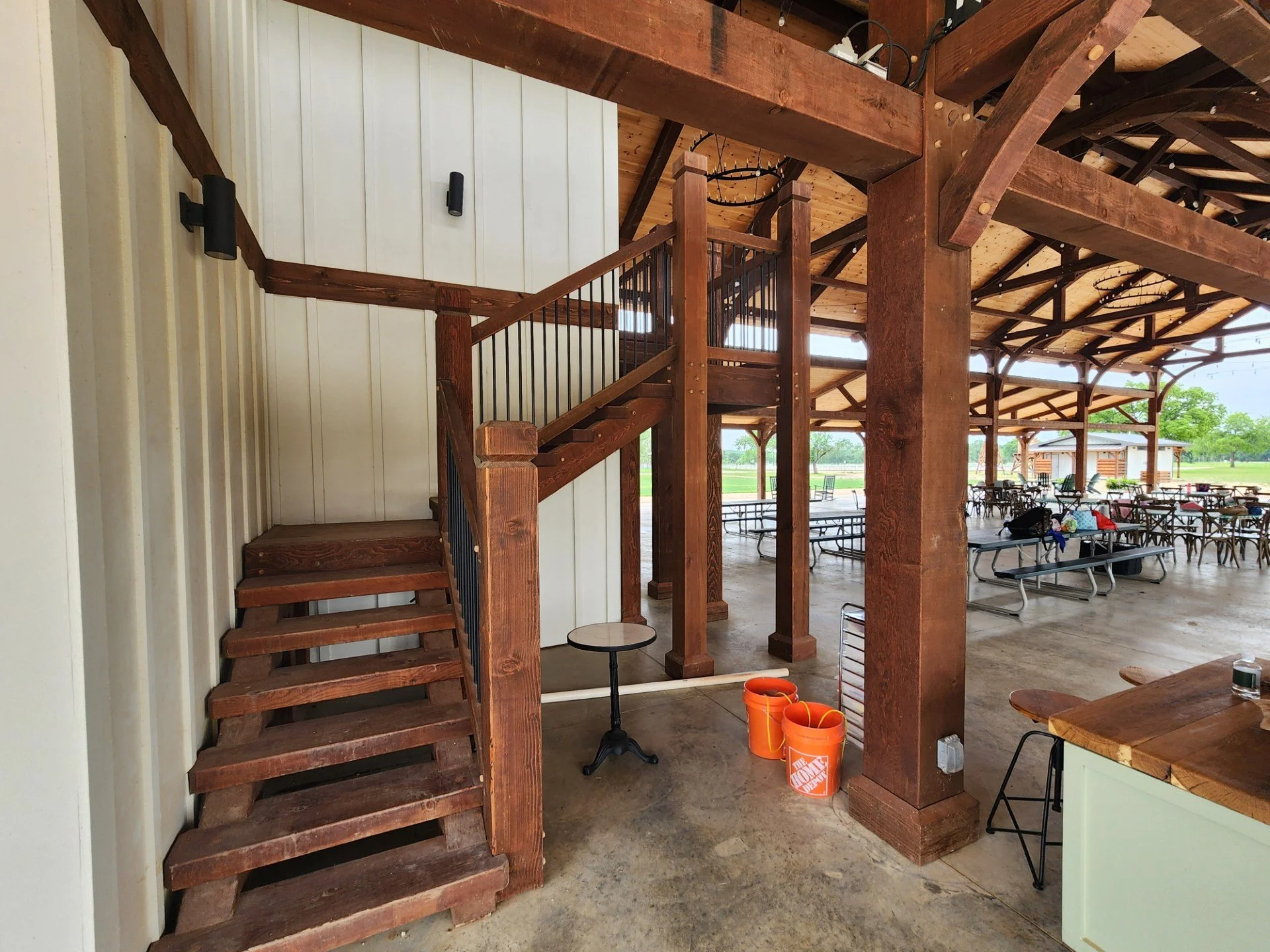 Wooden staircase next to a white paneled wall, leading up to a second level with wooden beams and rustic ceiling. Outdoor seating with tables and chairs in a covered area outdoors.