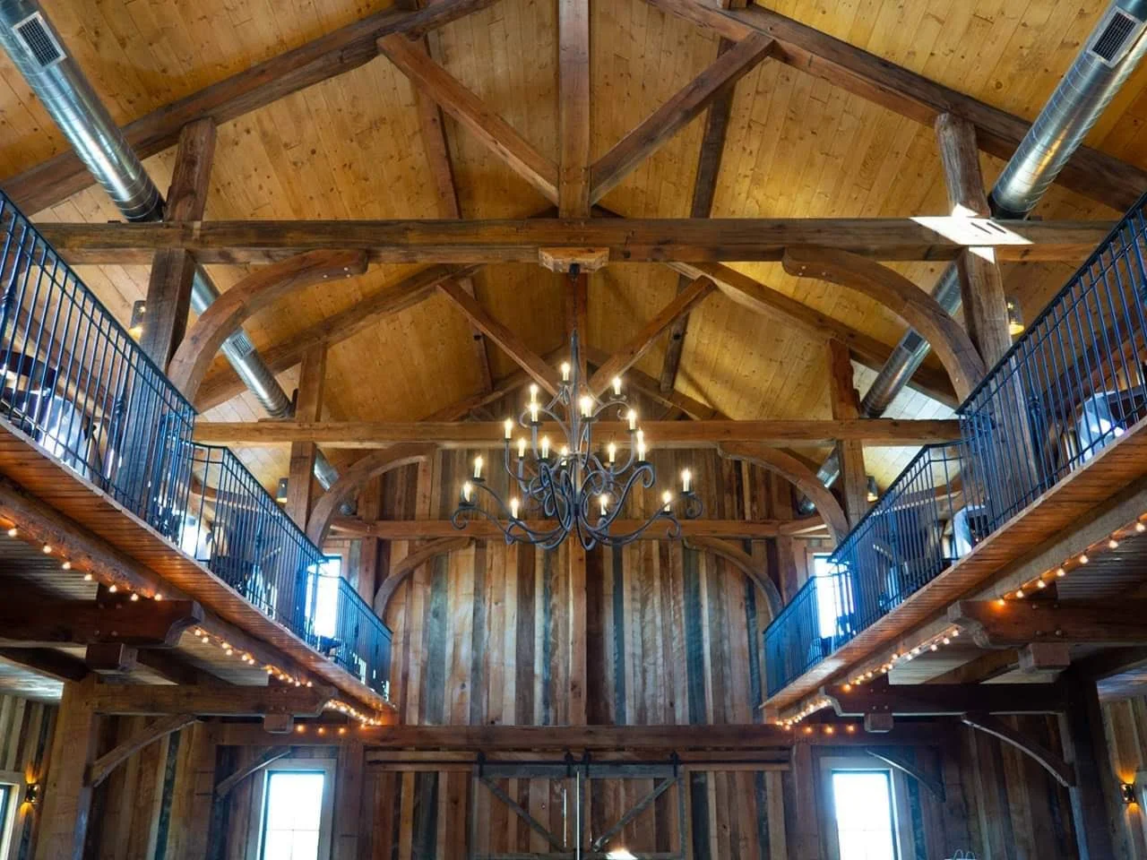 Interior of a wooden hall with high vaulted ceiling, a chandelier hanging from the center, and balcony railings along the sides with string lights.
