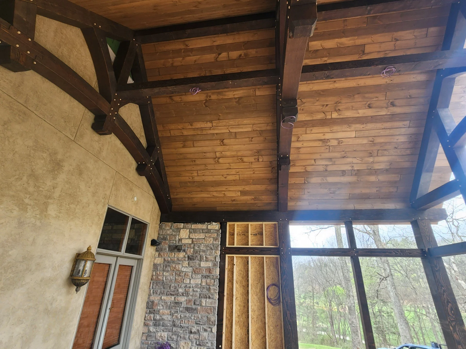 Construction of a screened porch or sunroom with a wooden ceiling and framing, brick wall, and sliding glass door, overlooking a wooded outdoor area.
