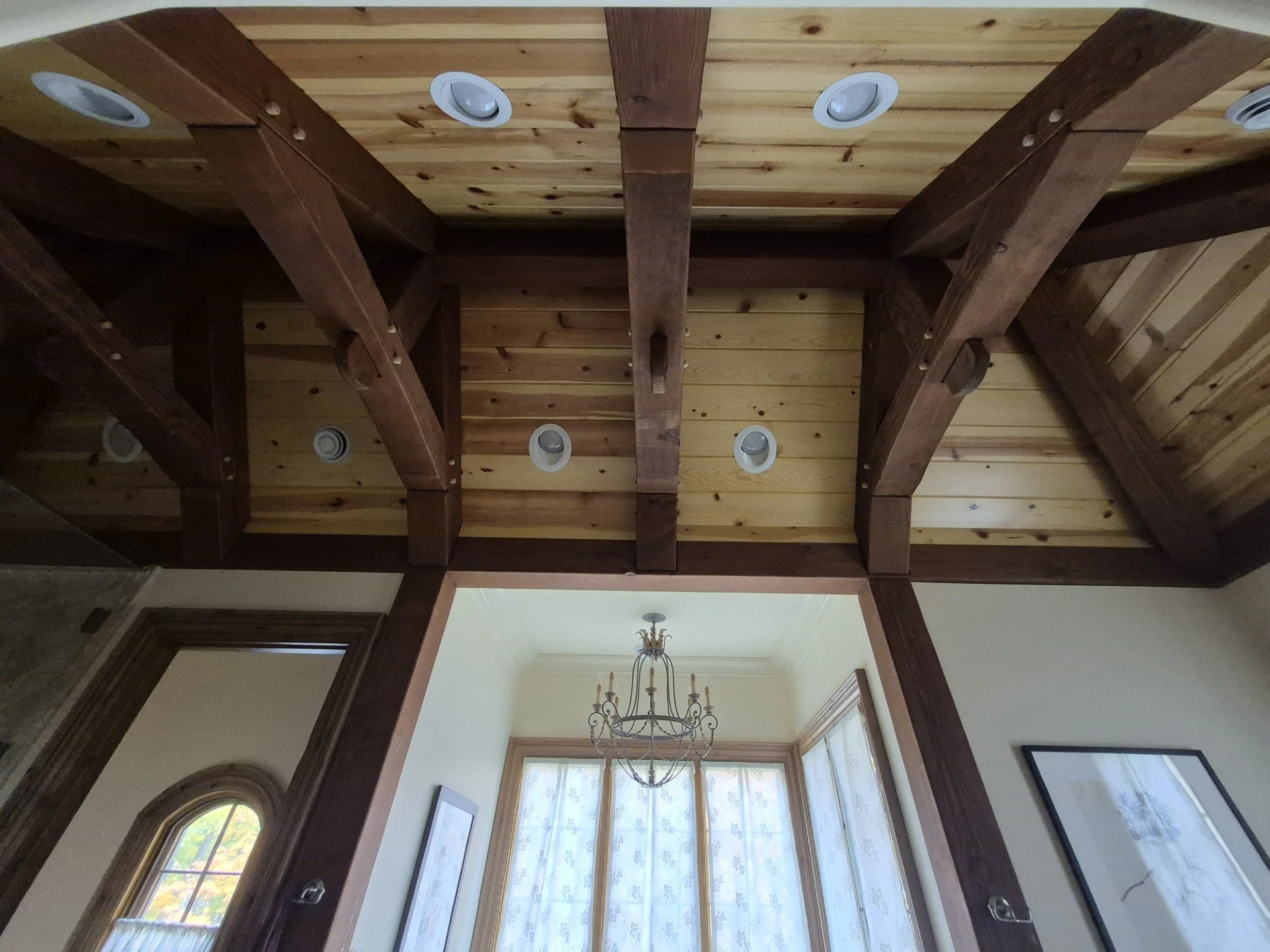 View of a wooden ceiling with exposed beams and recessed ceiling lights. A chandelier hangs below in front of a window with sheer curtains, adjacent to a wall with framed pictures.