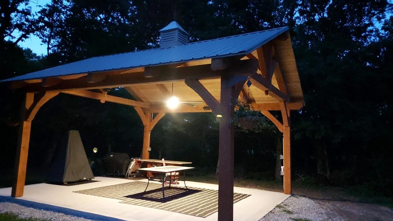An outdoor wooden pavilion with a metal roof illuminated by a hanging light, situated on a concrete patio with a striped outdoor rug, a picnic table, a grill, and hanging flower basket, surrounded by trees at dusk.