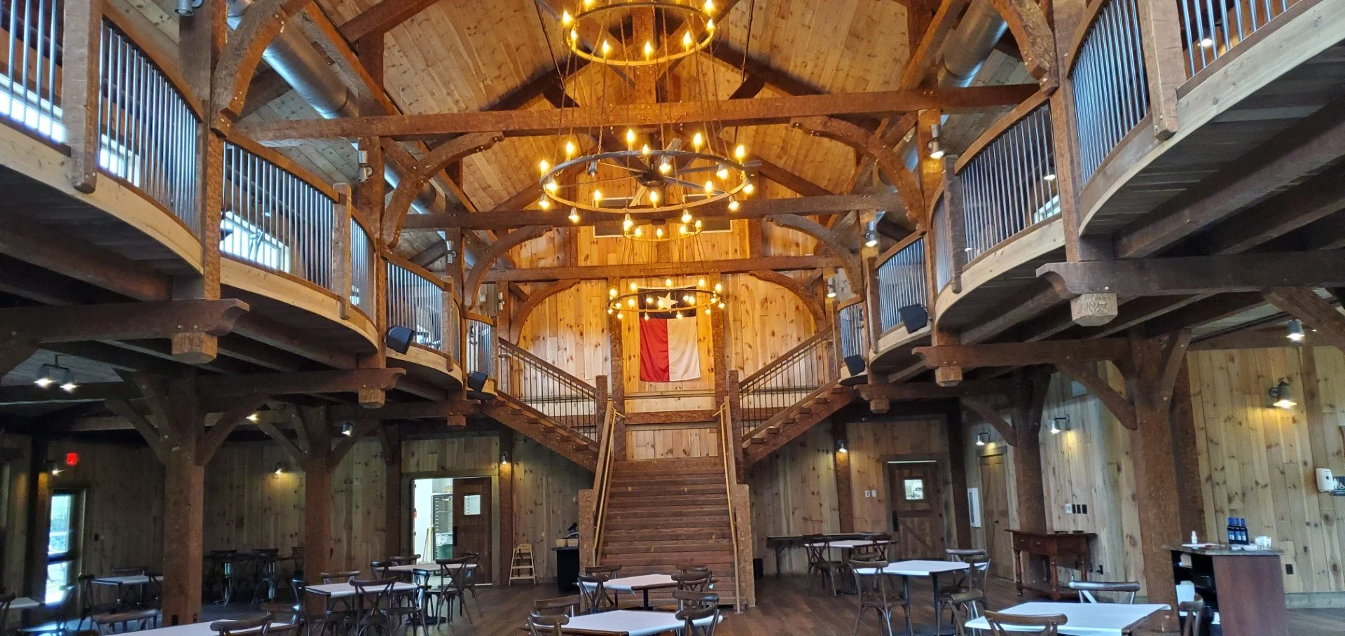 Interior of a rustic wooden hall with a high vaulted ceiling, circular chandeliers on ring frames, staircase at center, and a wall hanging of the American flag.
