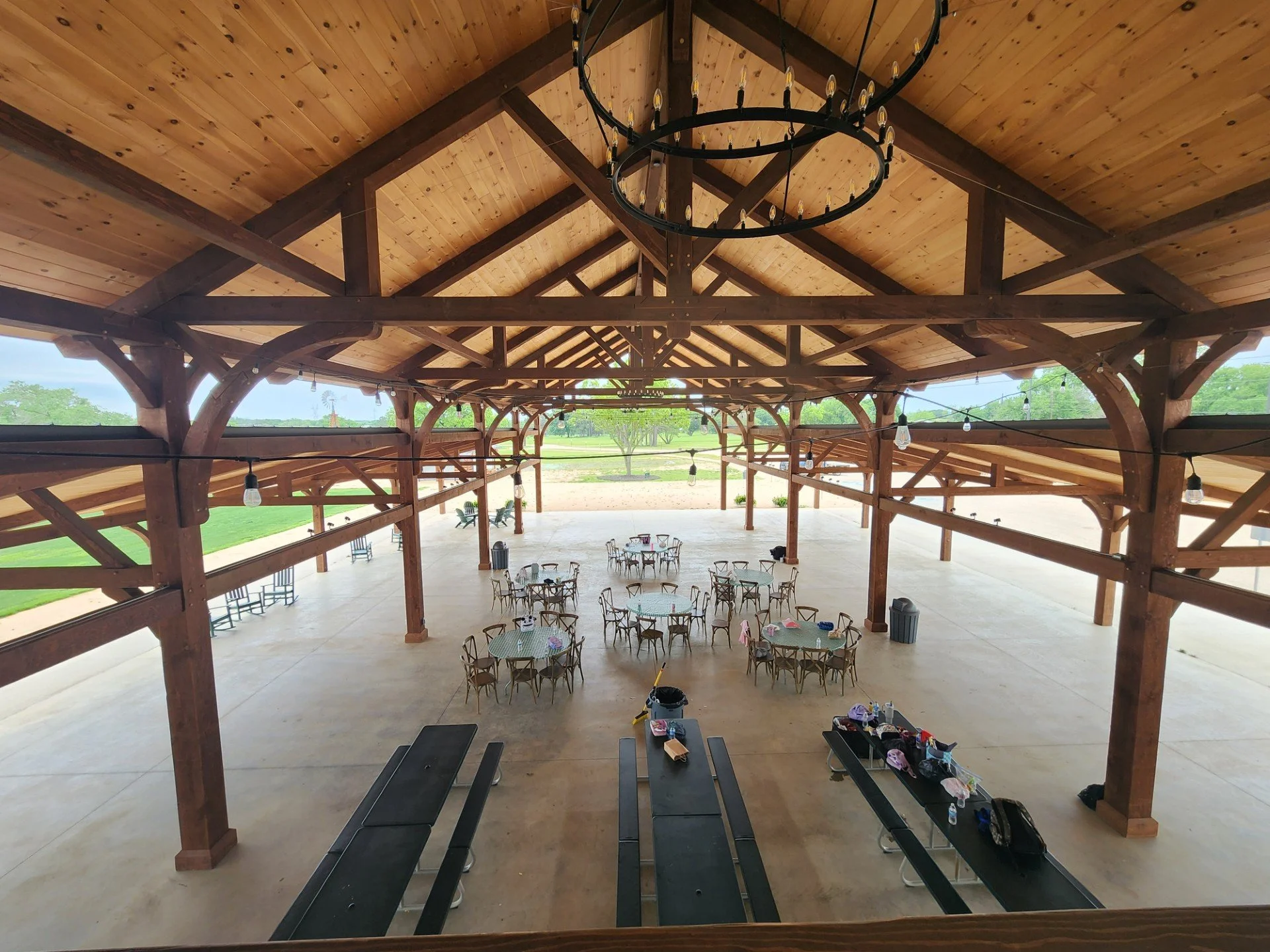 Inside of a wooden pavilion with a high ceiling, hanging circular chandeliers, string lights, and multiple round tables with chairs, set for an event.