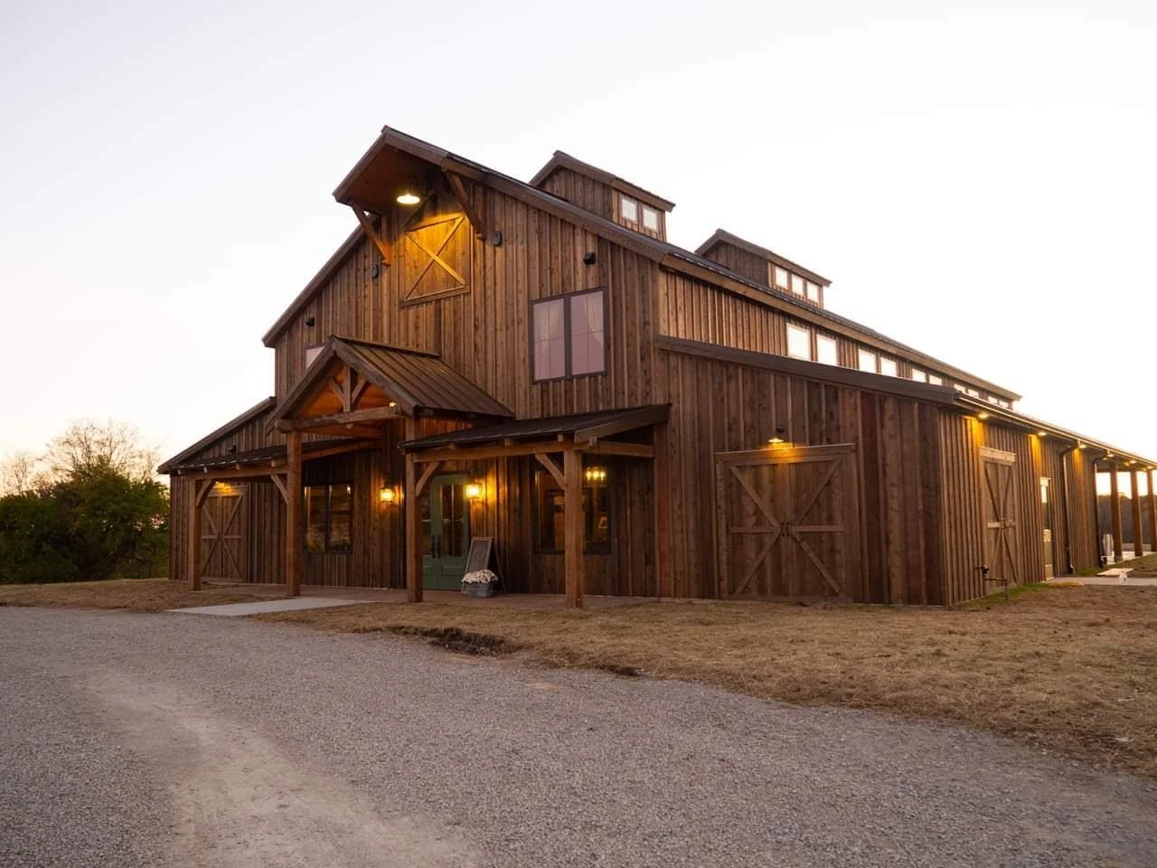 A large rustic wooden barn-style building with warm outdoor lighting, situated on a gravel driveway with dry grass surrounding it during sunset.