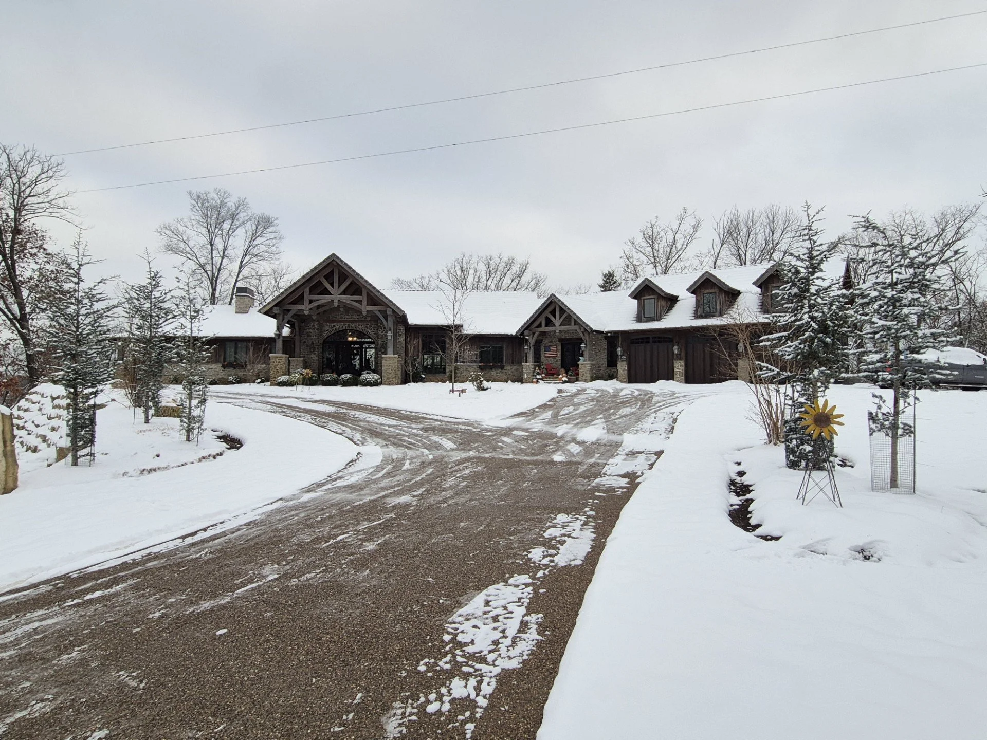 A large house in a snowy landscape with a curved gravel driveway, surrounded by snow-covered trees and shrubs, and a cloudy sky overhead.