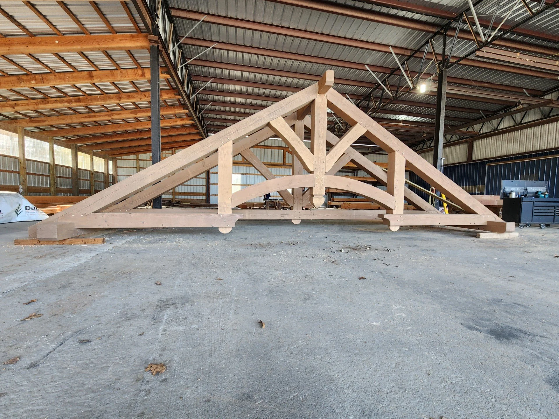 Wooden framework for a bridge or similar structure located inside a large industrial warehouse or workshop with a high metal roof and supporting beams.