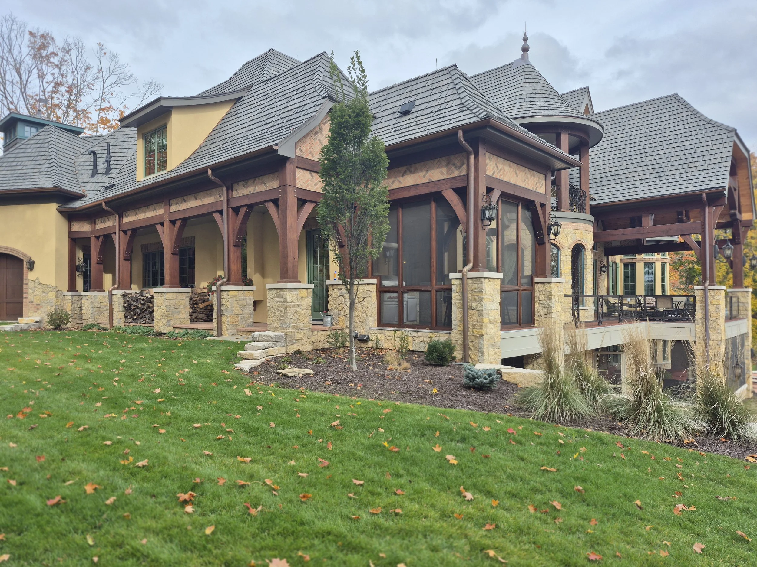 A large house with a complex roof structure, wooden beams, stone and stucco exterior, and a small front yard with grass and fallen leaves.