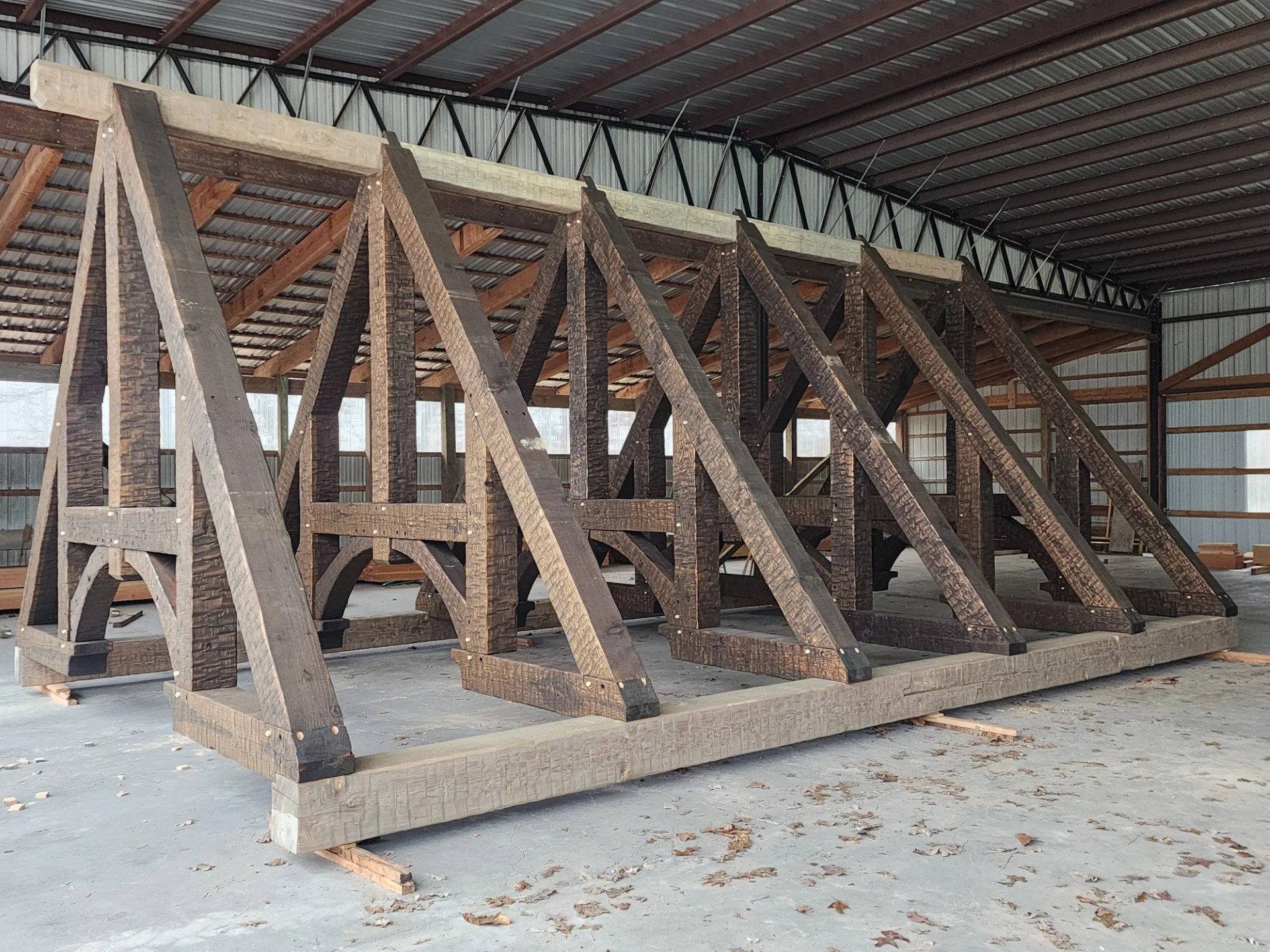 Interior of a barn under construction with wooden trusses and a metal roof, scattered leaves on the concrete floor.