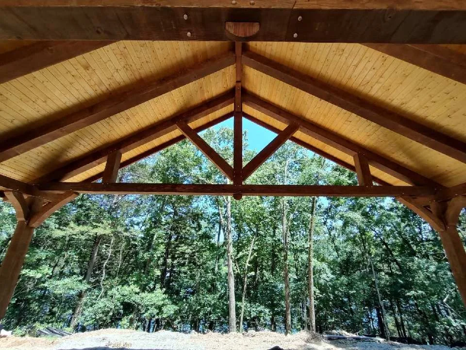 Wooden pavilion with a triangular roof, supported by wooden beams, overlooking a forest with tall green trees.