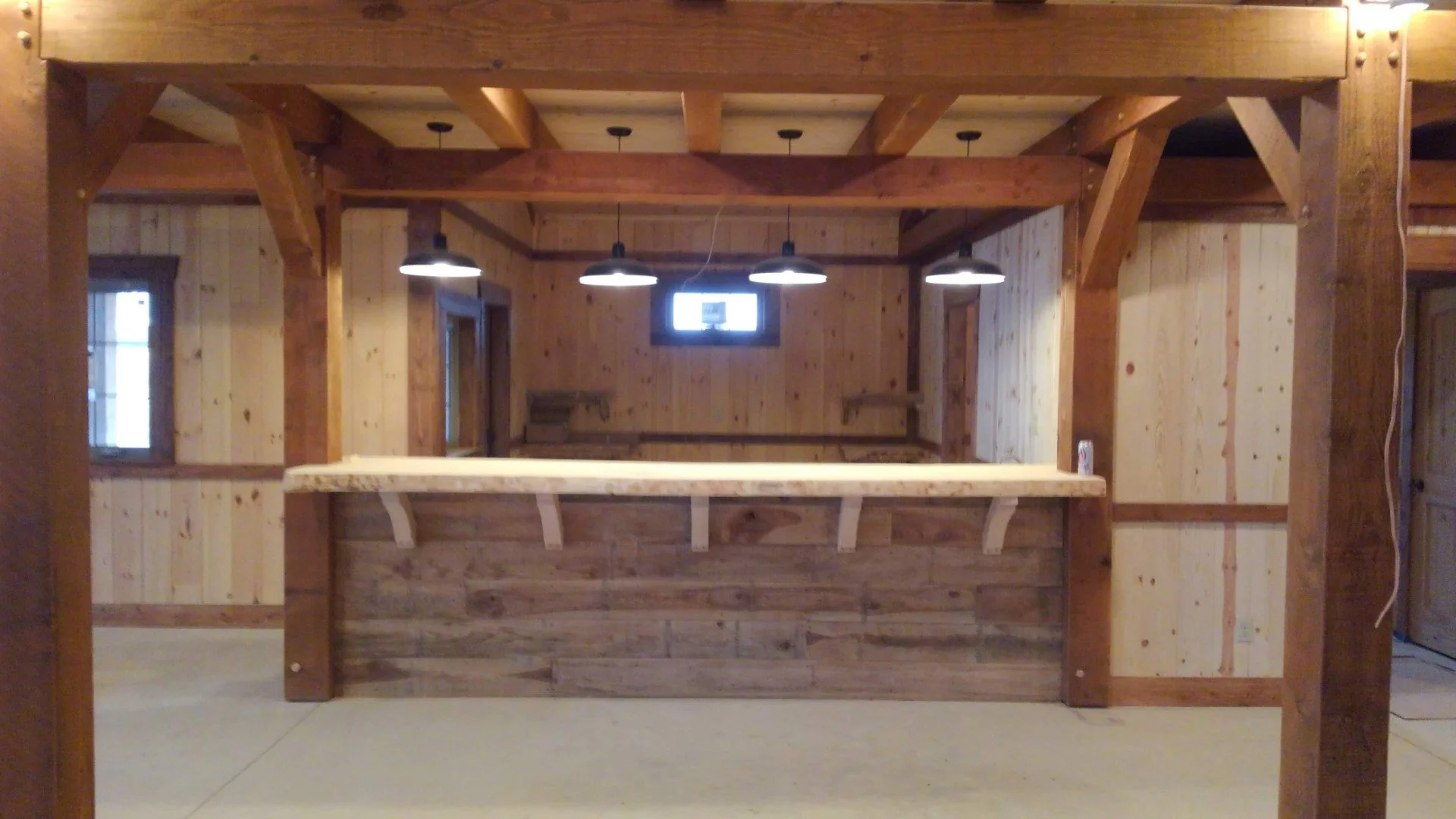 Interior view of a rustic wooden cabin bar with a wooden countertop and wooden walls. Three black pendant lights hanging from the ceiling, and a small window in the back wall.