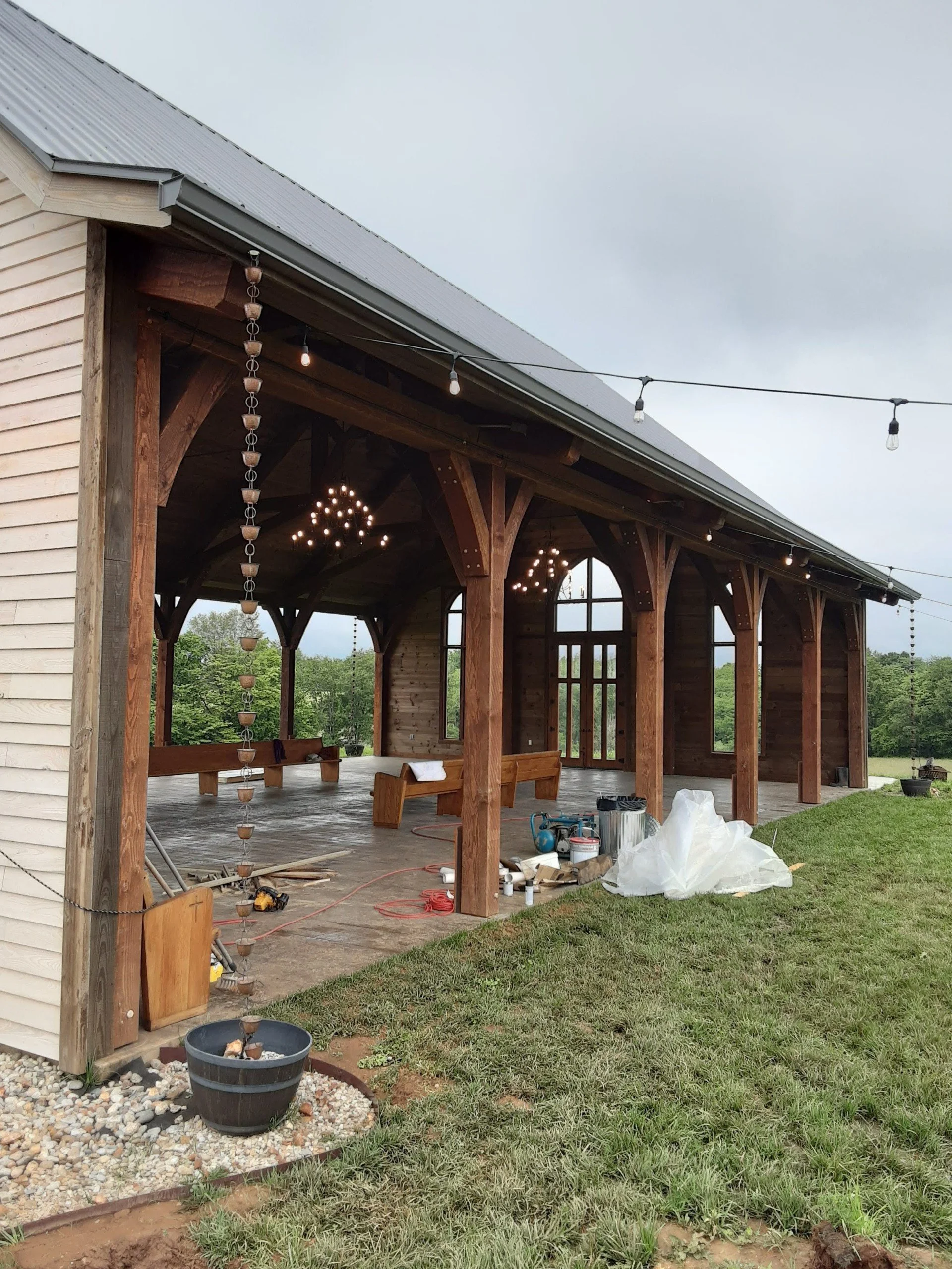 Under construction patio or outdoor living area with wooden beams, string lights, and hanging strings of small planters or lights, surrounded by grass and a partially finished building with white siding and a metal roof.