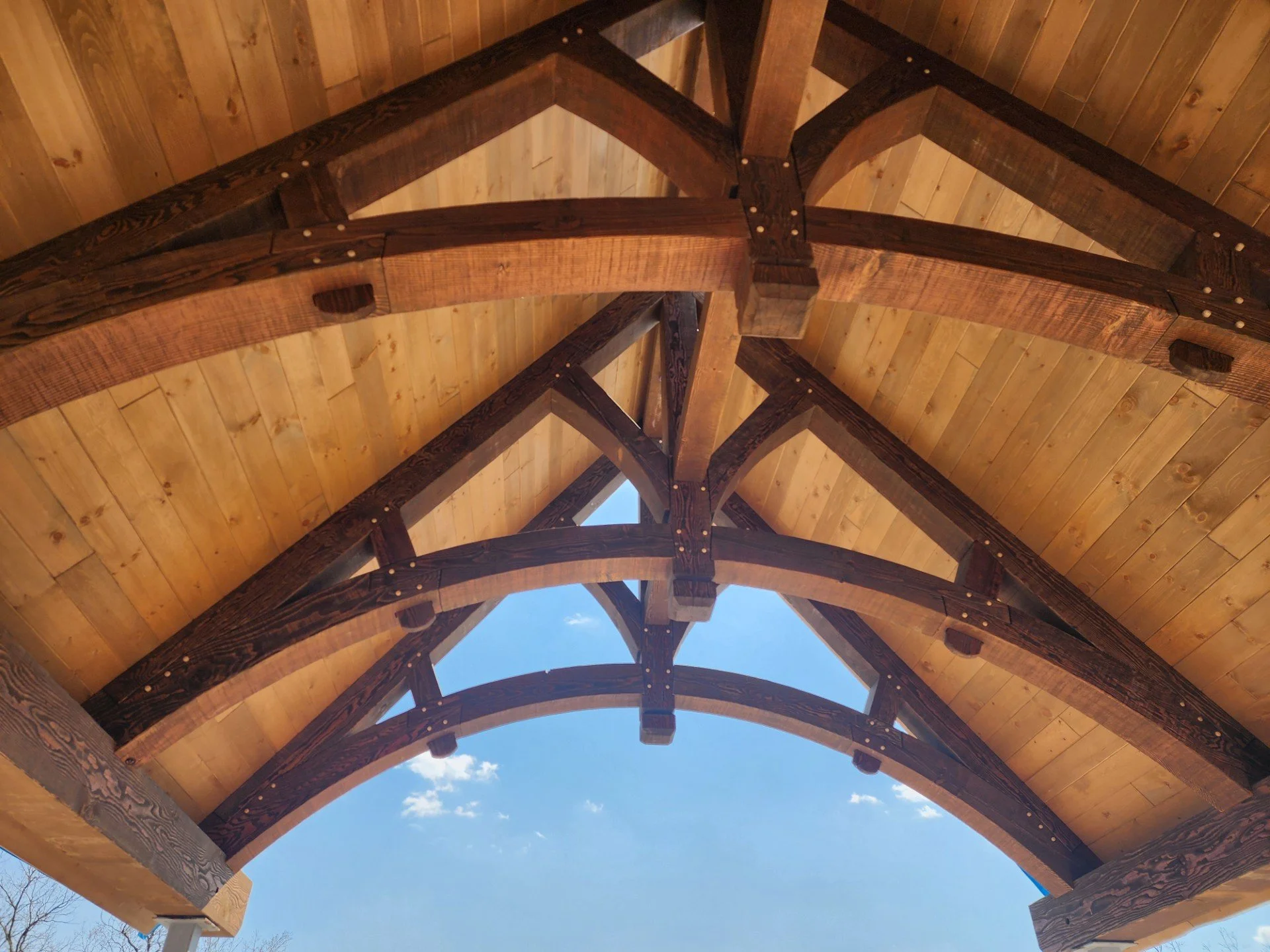 Looking up at a wooden construction with curved dark timber beams and lighter wood panels, showing a partly cloudy sky through the gaps.