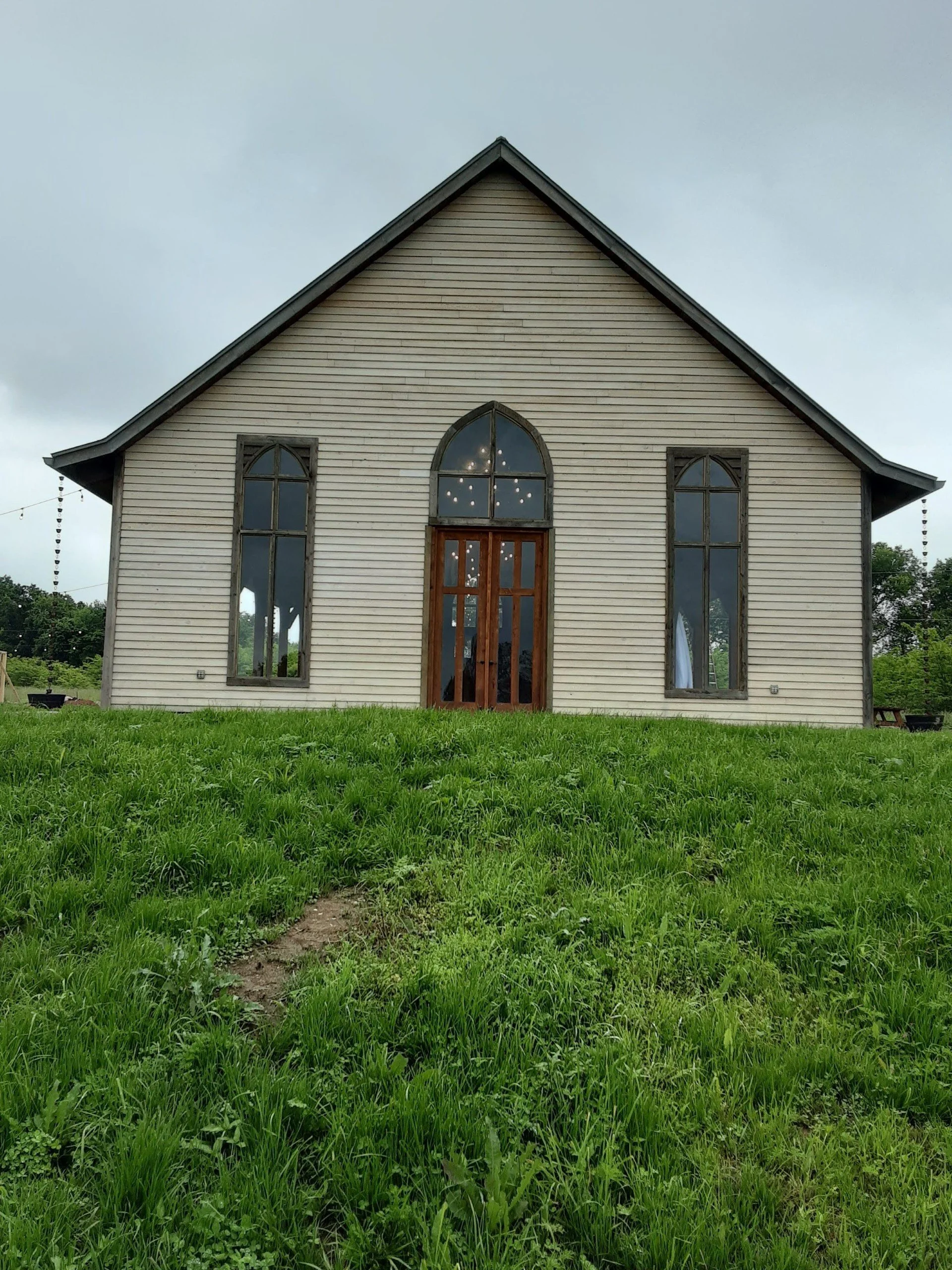 A small beige church with a wooden door and three tall, narrow arched windows on a grassy field, cloudy sky in the background.