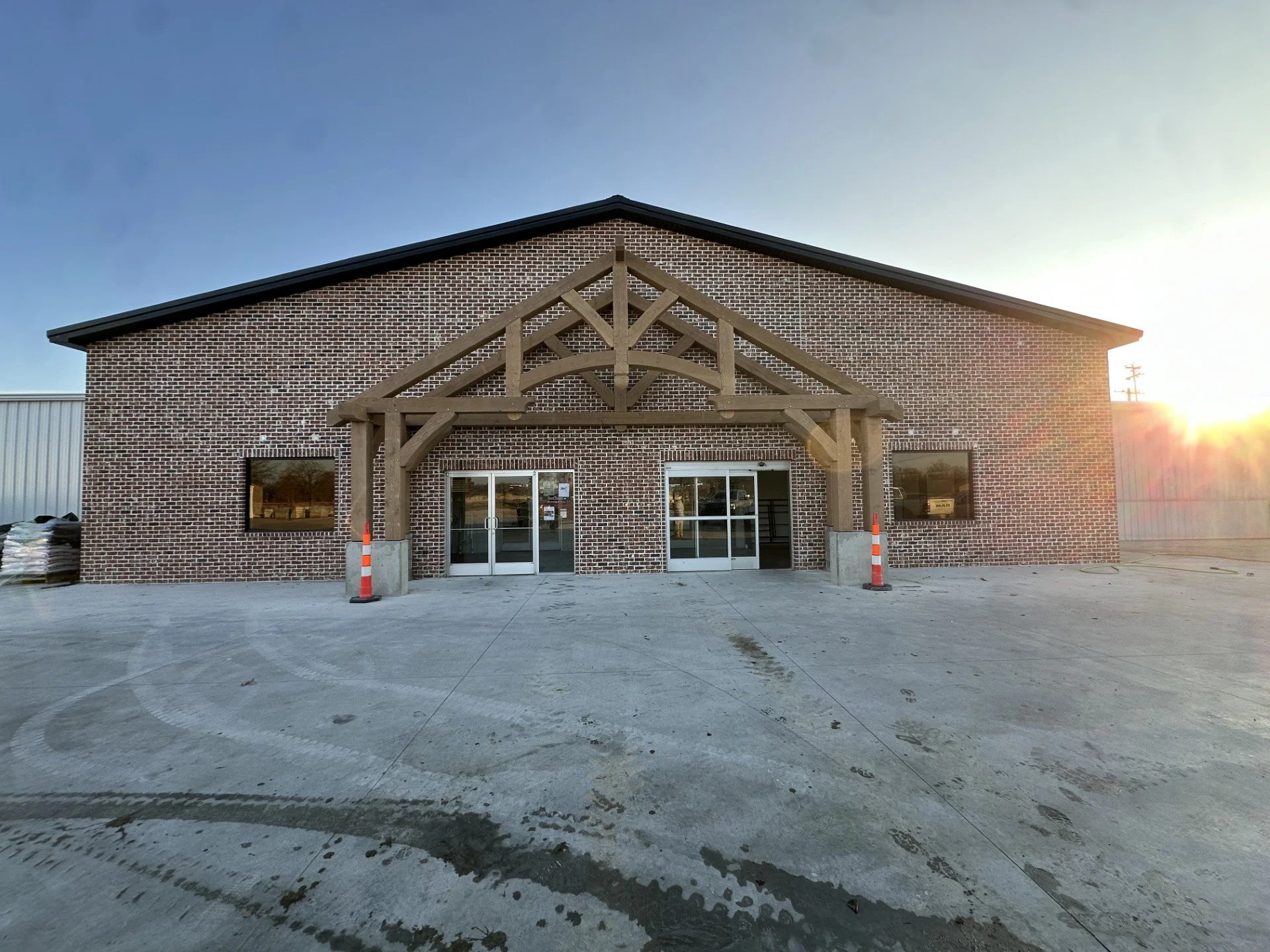 Front of a brick building with a peaked roof and decorative wooden beams above the entrance, with glass double doors, flanked by two windows, and orange and white traffic cones on either side. The ground has tire marks and a slight wet stain, and the