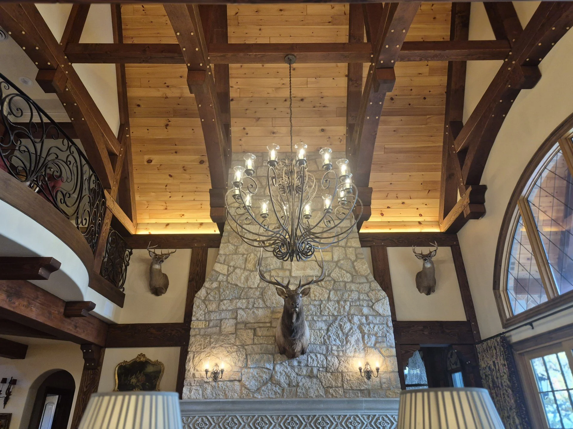 Interior view of a rustic living room with a high ceiling, showcasing dark wood beams, a large stone fireplace with a mounted deer head, and two mounted deer heads on either side of the fireplace. There is an ornate chandelier hanging from the ceilin