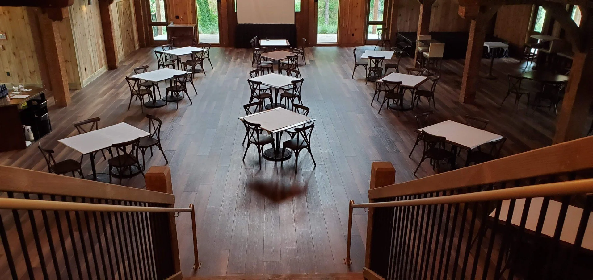 An empty dining hall with wooden walls and floors, several square tables with white tablecloths, and black chairs. Large windows let in natural light, and there are doors at the back leading outside.