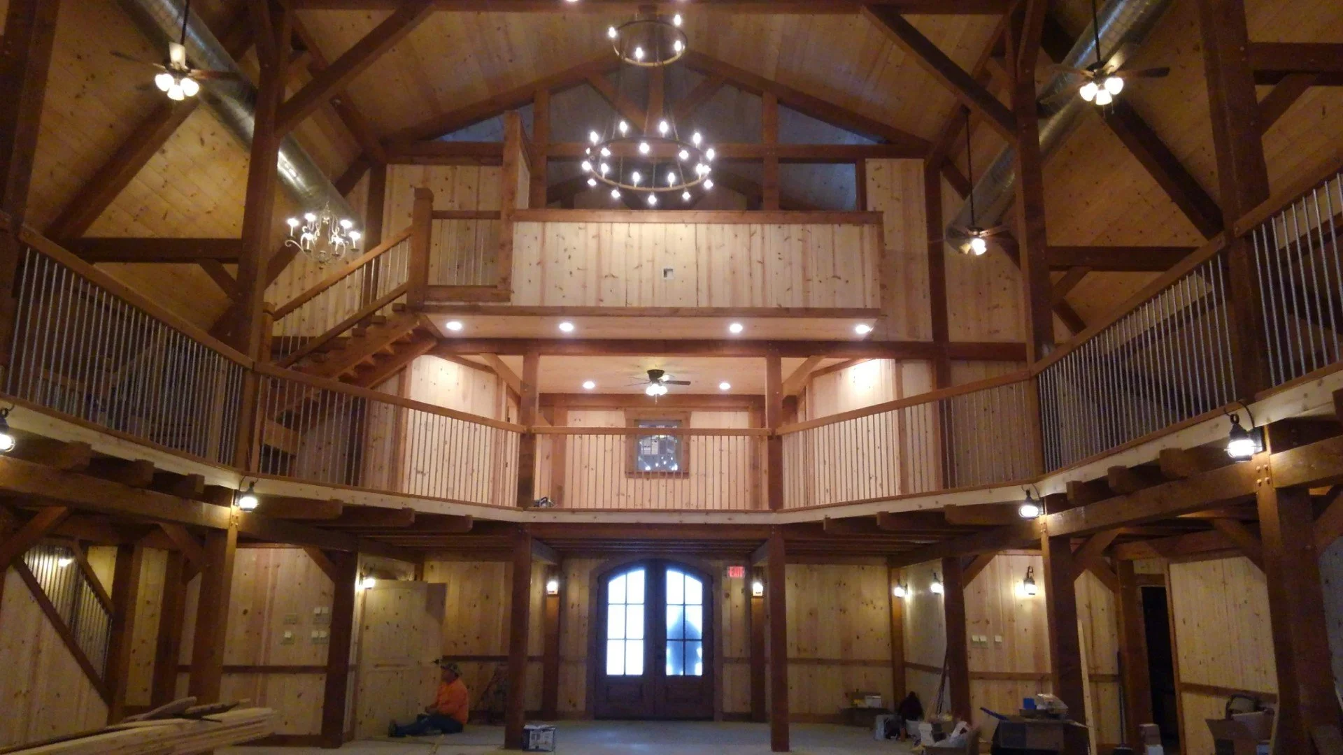 Interior view of a large wooden building with second-floor balcony, high vaulted ceiling, ceiling fans, and hanging lights, under construction.