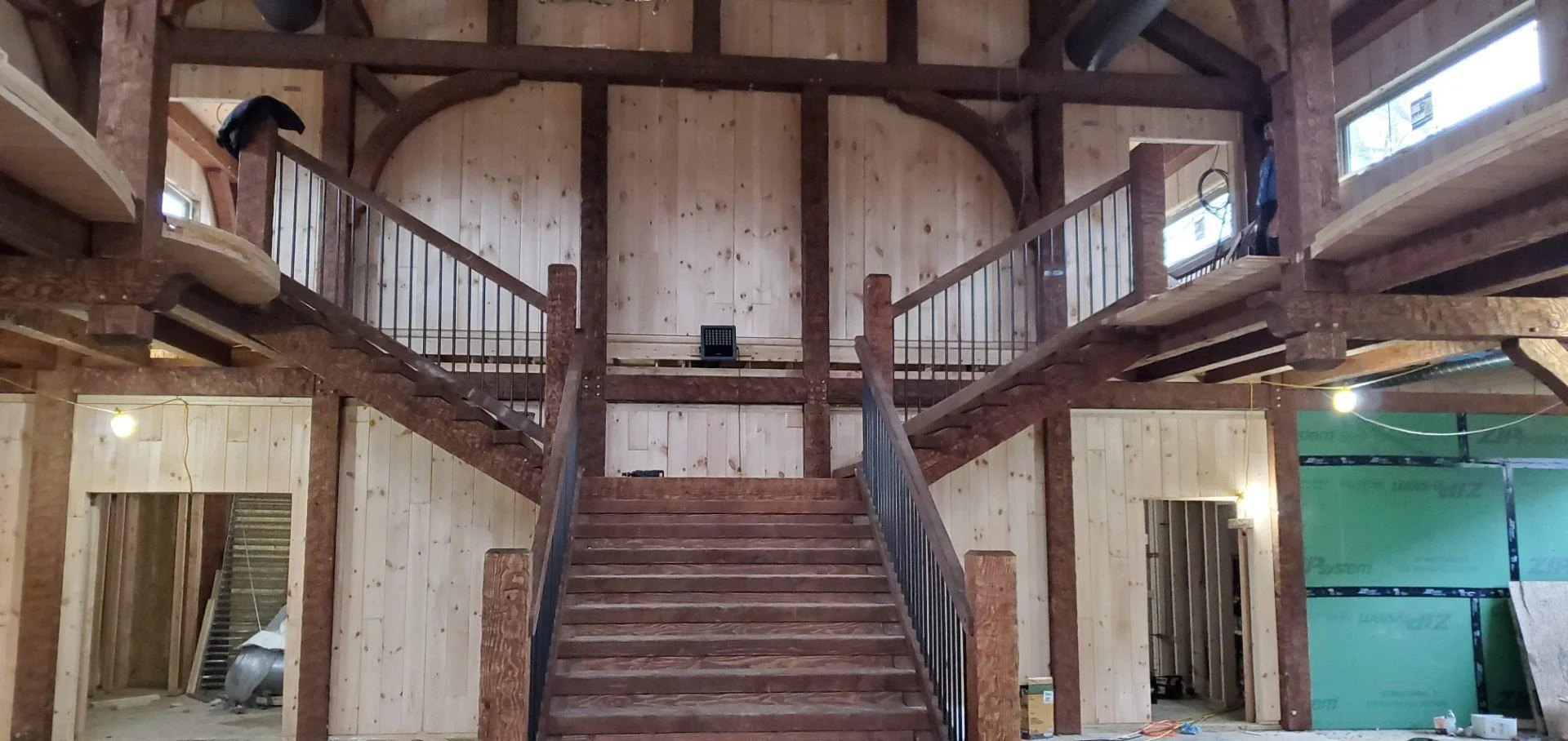 Interior view of a house under construction with a wooden staircase leading to a second floor. The space features exposed wooden beams and walls, with construction materials and tools visible.