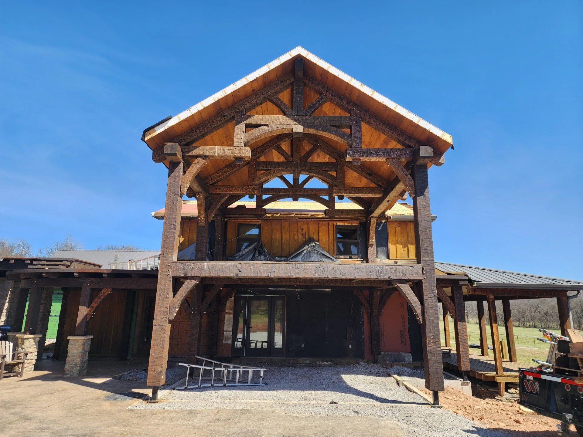 Construction site of a house with wooden beams and roof framework, partially built with open space and windows, under a clear blue sky.