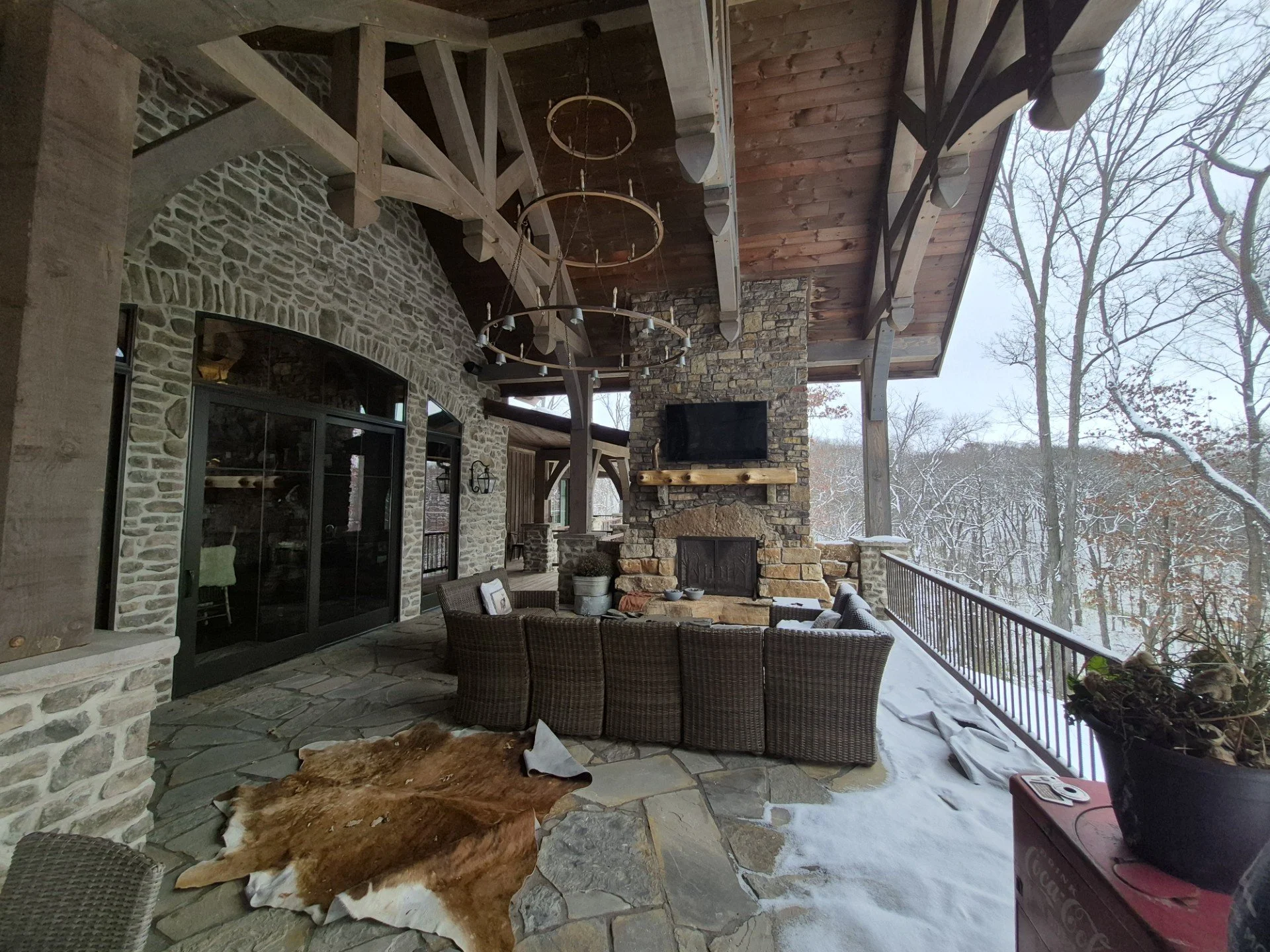 A covered outdoor patio area with stone flooring, a stone fireplace, a mounted TV above the fireplace, a wooden mantel, a wicker outdoor sofa, a cowhide rug, and patio furniture. Snow is visible outside the railing, and leafless trees are in the back