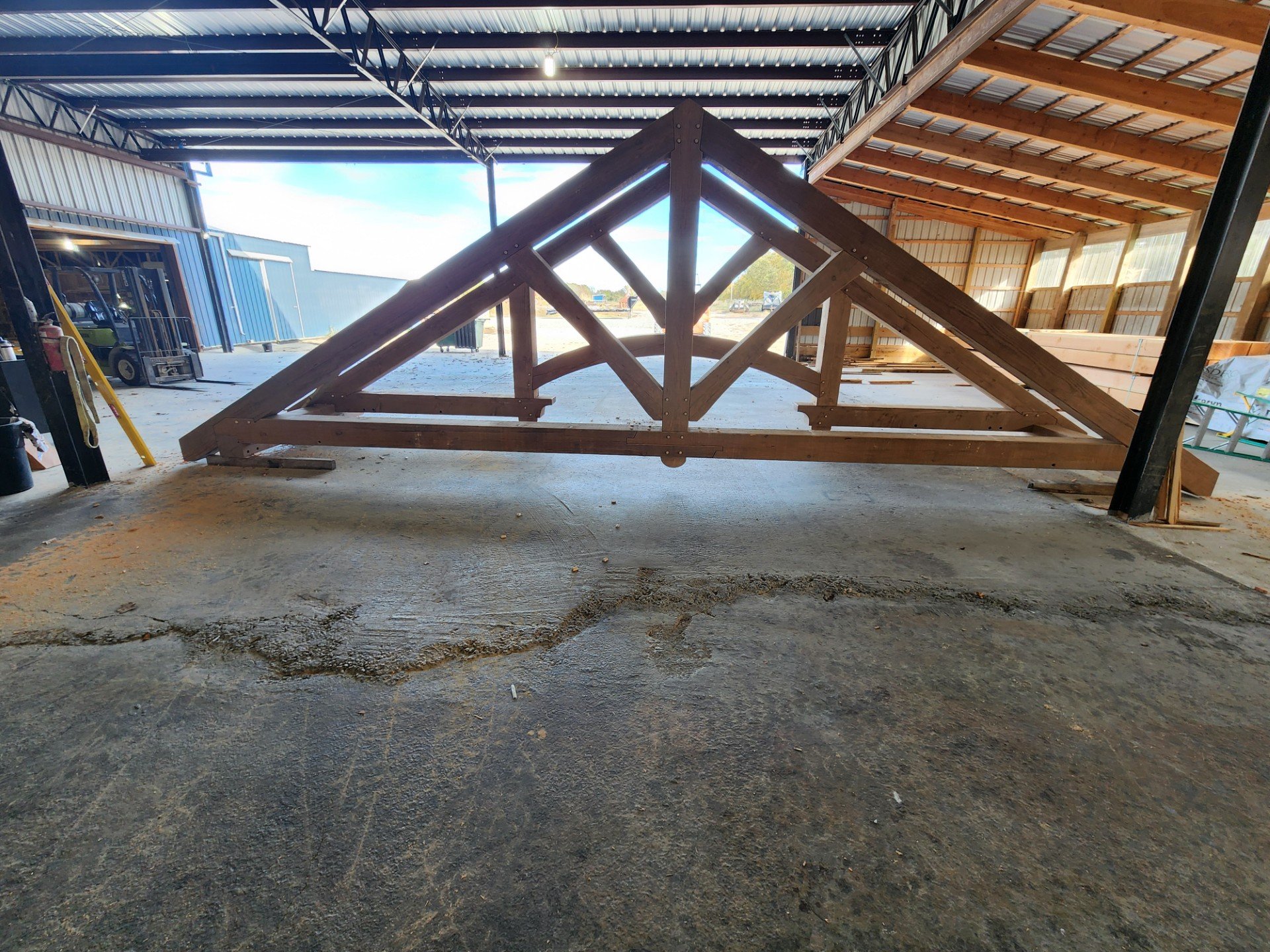 Indoor construction site showing wooden framing structure on a concrete floor, with a partially built roof and an outside view through the open side of the building.