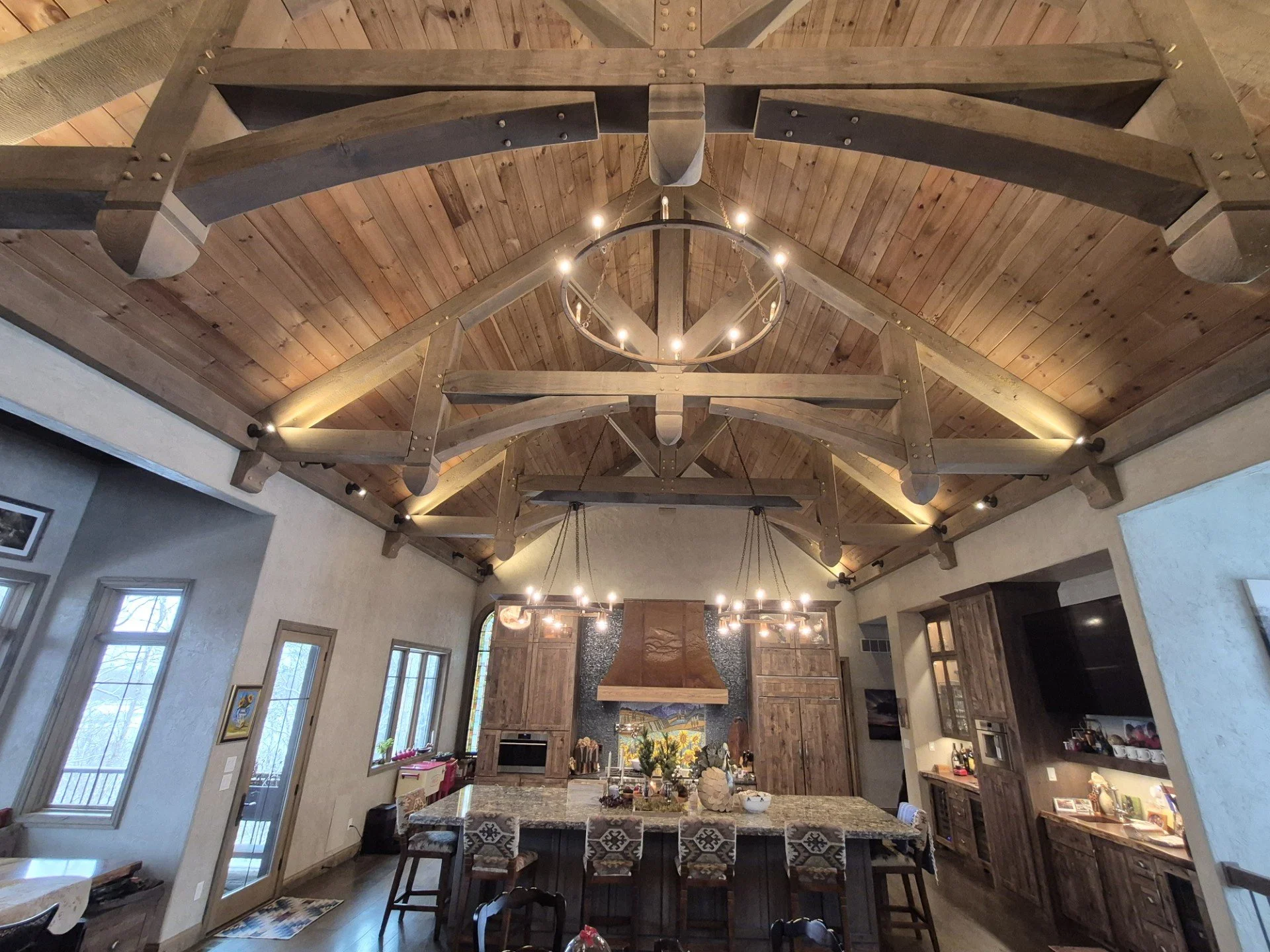 Open-concept kitchen with a large island, wooden cabinets, and high vaulted wooden ceilings featuring large wooden beams and circular chandeliers.