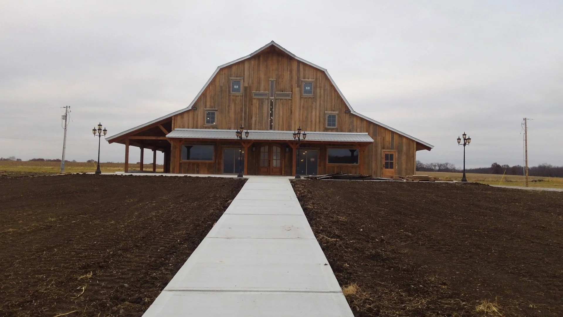 A rustic wooden church with a cross on the front, surrounded by open land and a straight concrete walkway leading to the entrance, under an overcast sky.