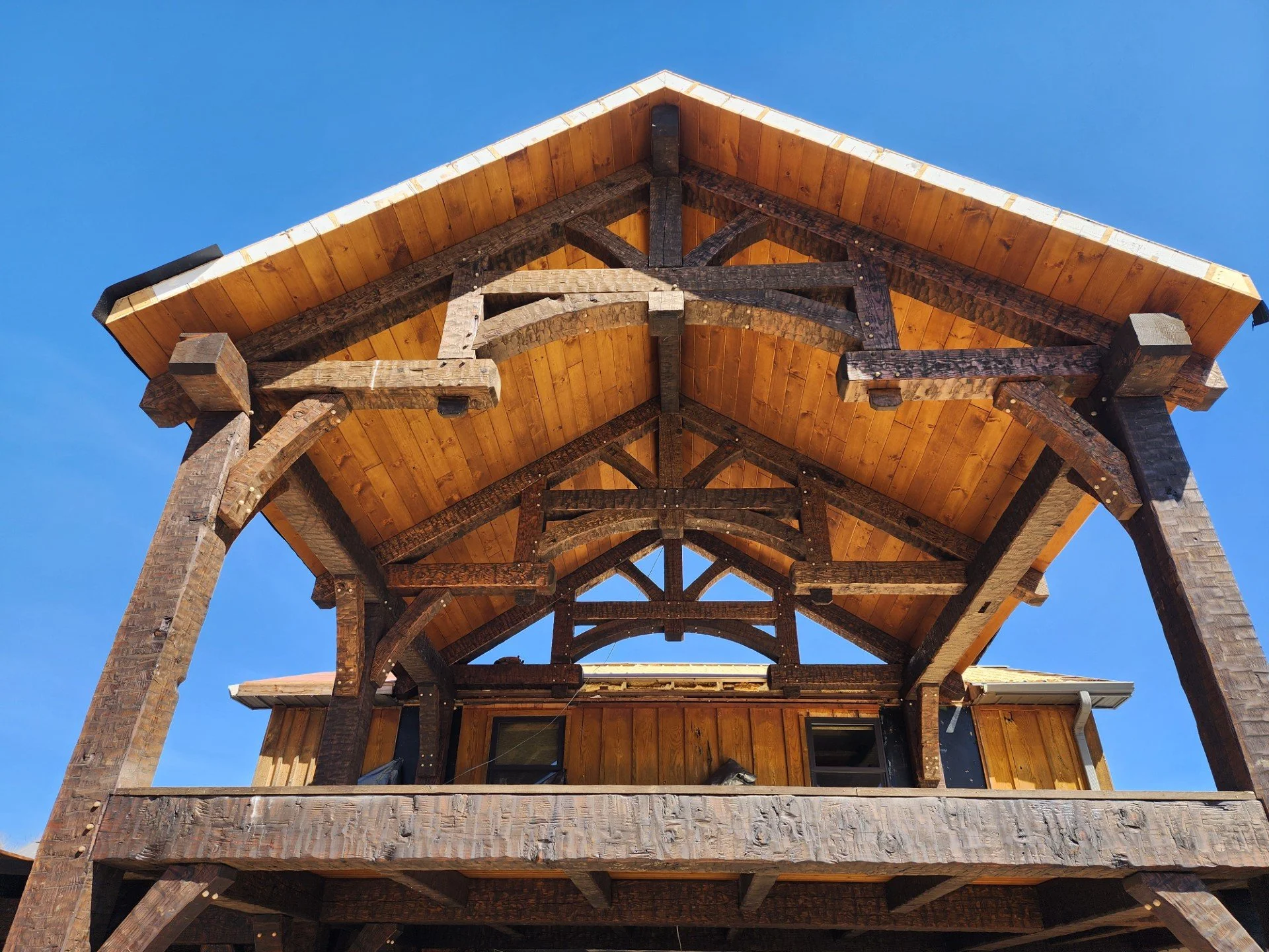 View of a wooden house under construction, showing the roof and part of the upper floor framework against a clear blue sky.